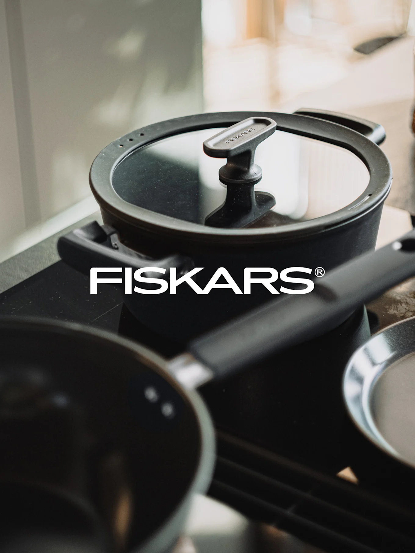 Une casserole noire avec un couvercle en verre et le logo blanc de Fiskars sur une cuisinière, avec une poêle floue au premier plan.