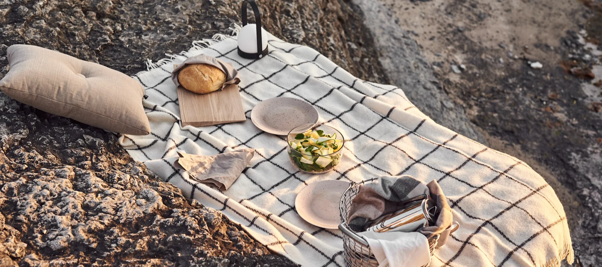 Un ensemble de pique-nique sur des rochers accidentés, avec une couverture à carreaux blanche, un coussin, du pain, un saladier, des assiettes, une lanterne et un panier avec une couverture.