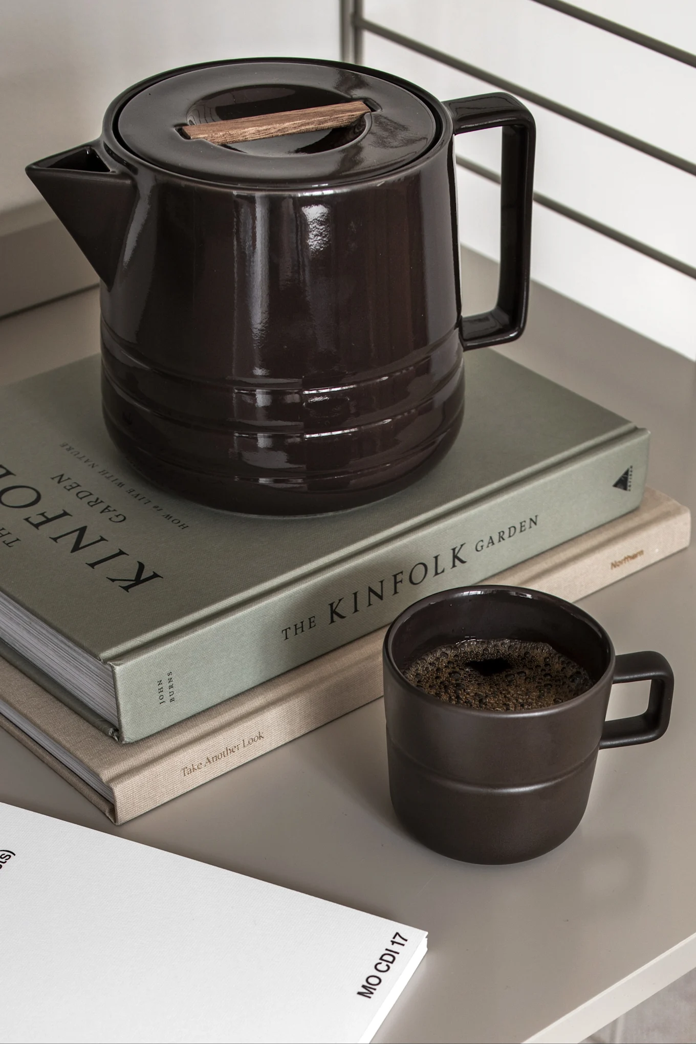 Cafetière en céramique marron foncé avec couvercle en bois, une tasse assortie avec du café et deux livres.