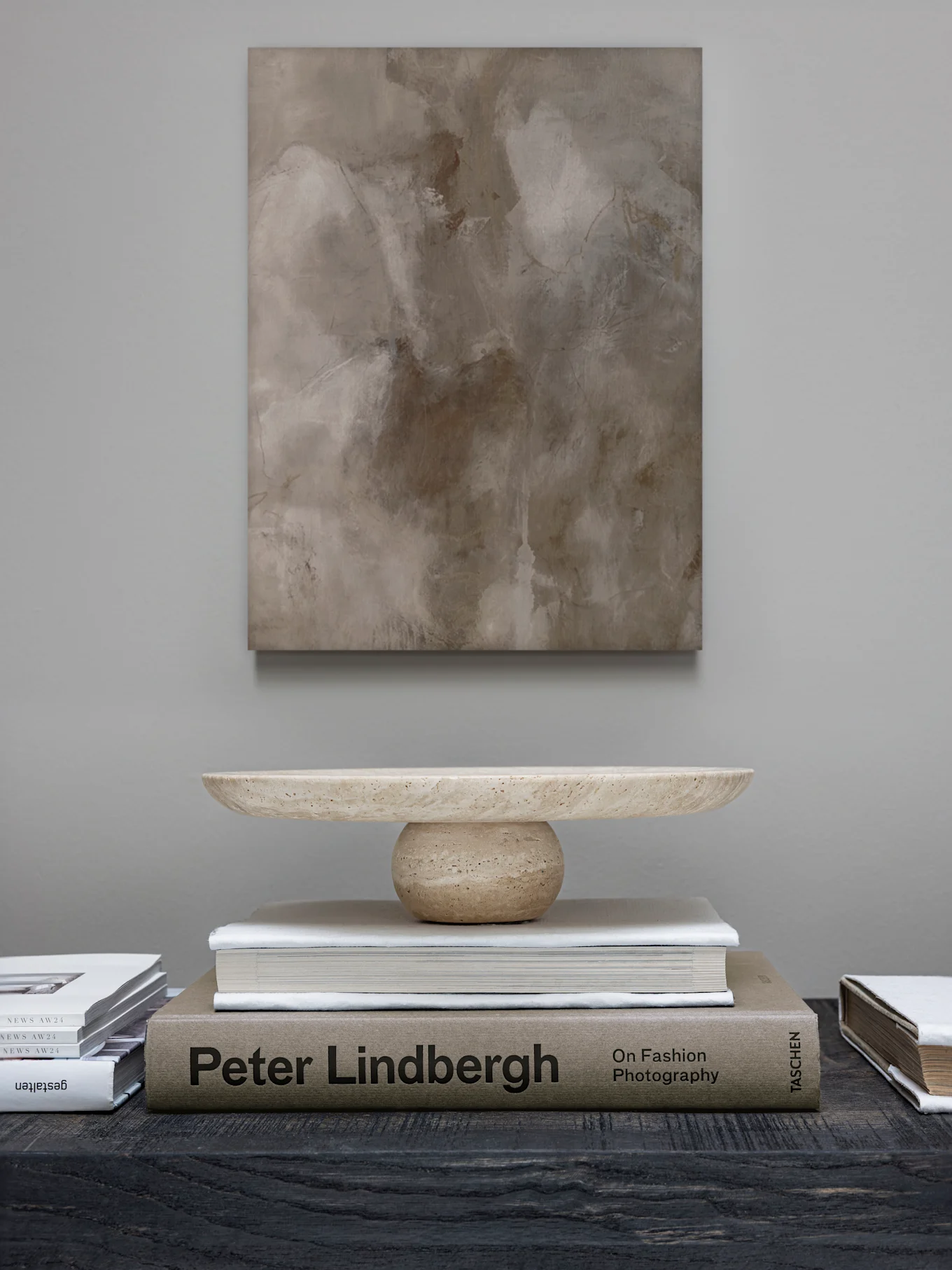 Une peinture abstraite est suspendue au-dessus d'un bol décoratif en pierre claire sur une pile de livres, dont 'Peter Lindbergh', sur une table sombre.