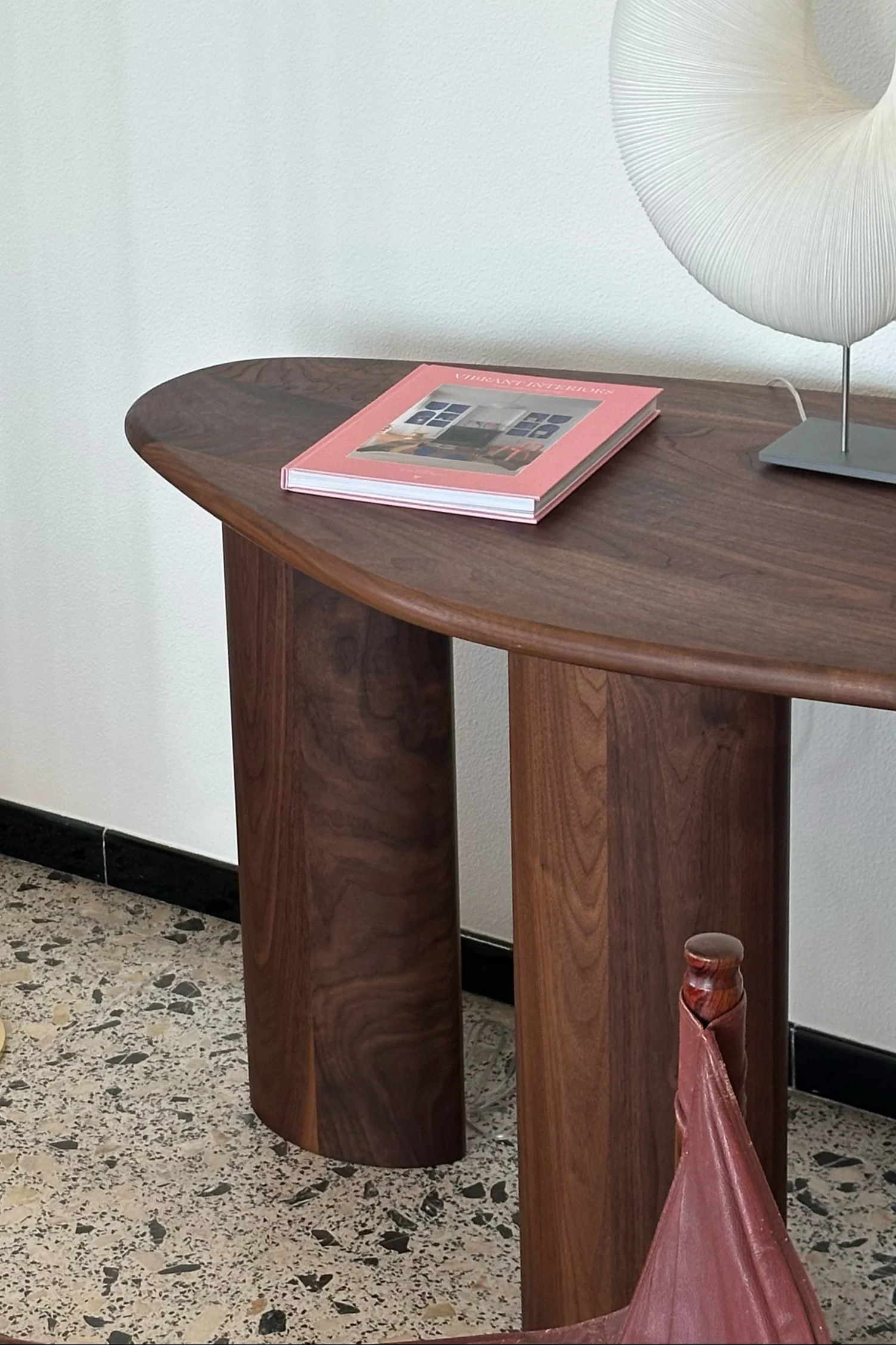 Une console moderne en bois foncé, avec un livre rose et une lampe blanche plissée, posée sur un sol en terrazzo.