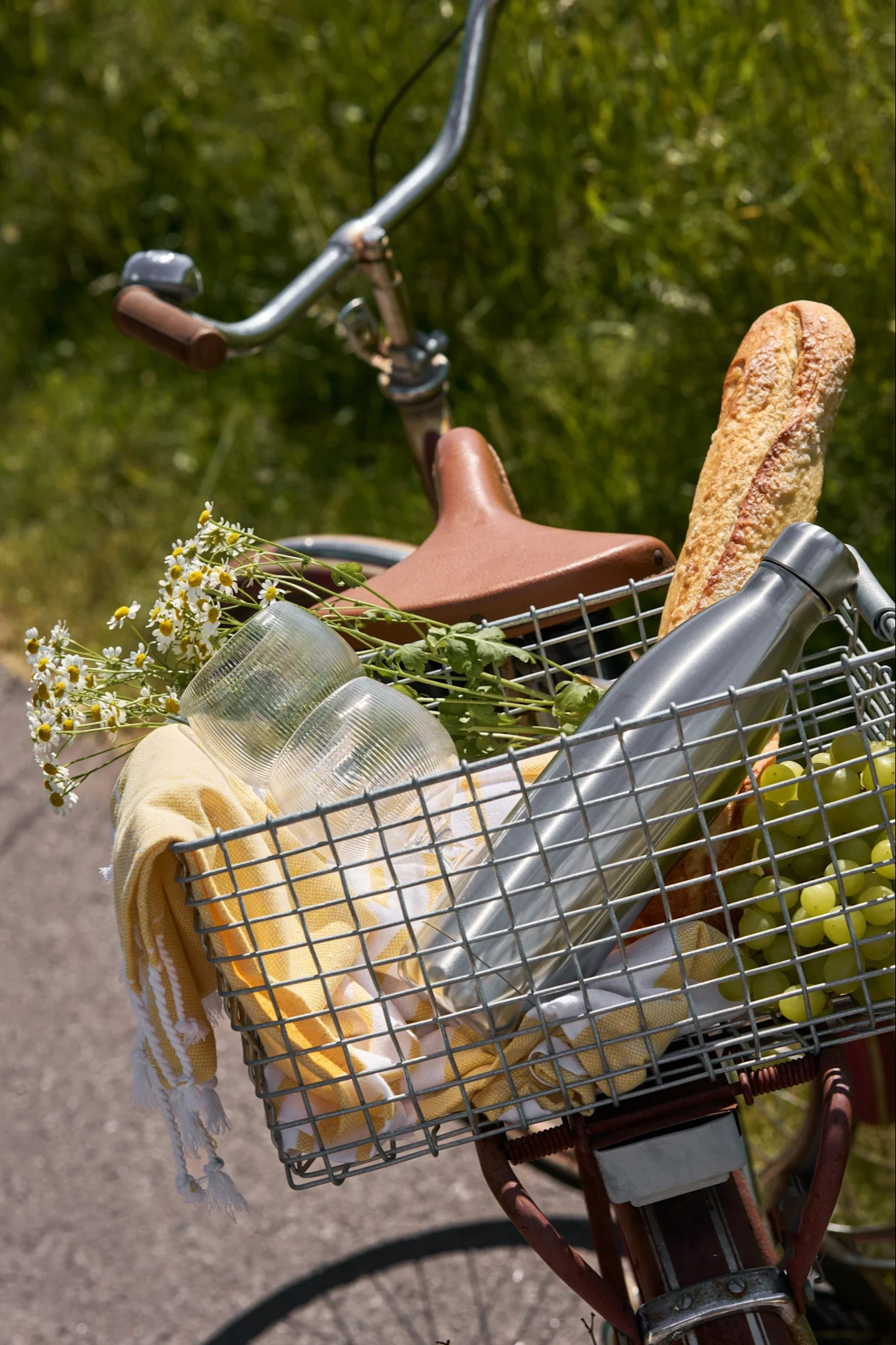 Panier avant de vélo rempli d'articles de pique-nique : une baguette, une bouteille d'eau, des raisins, des verres, une serviette jaune et des marguerites.