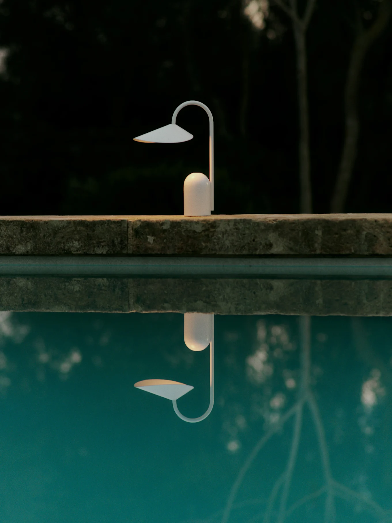 Lampe moderne blanche sur le bord d'une piscine, se reflétant dans l'eau bleue sur un fond naturel sombre et flou.