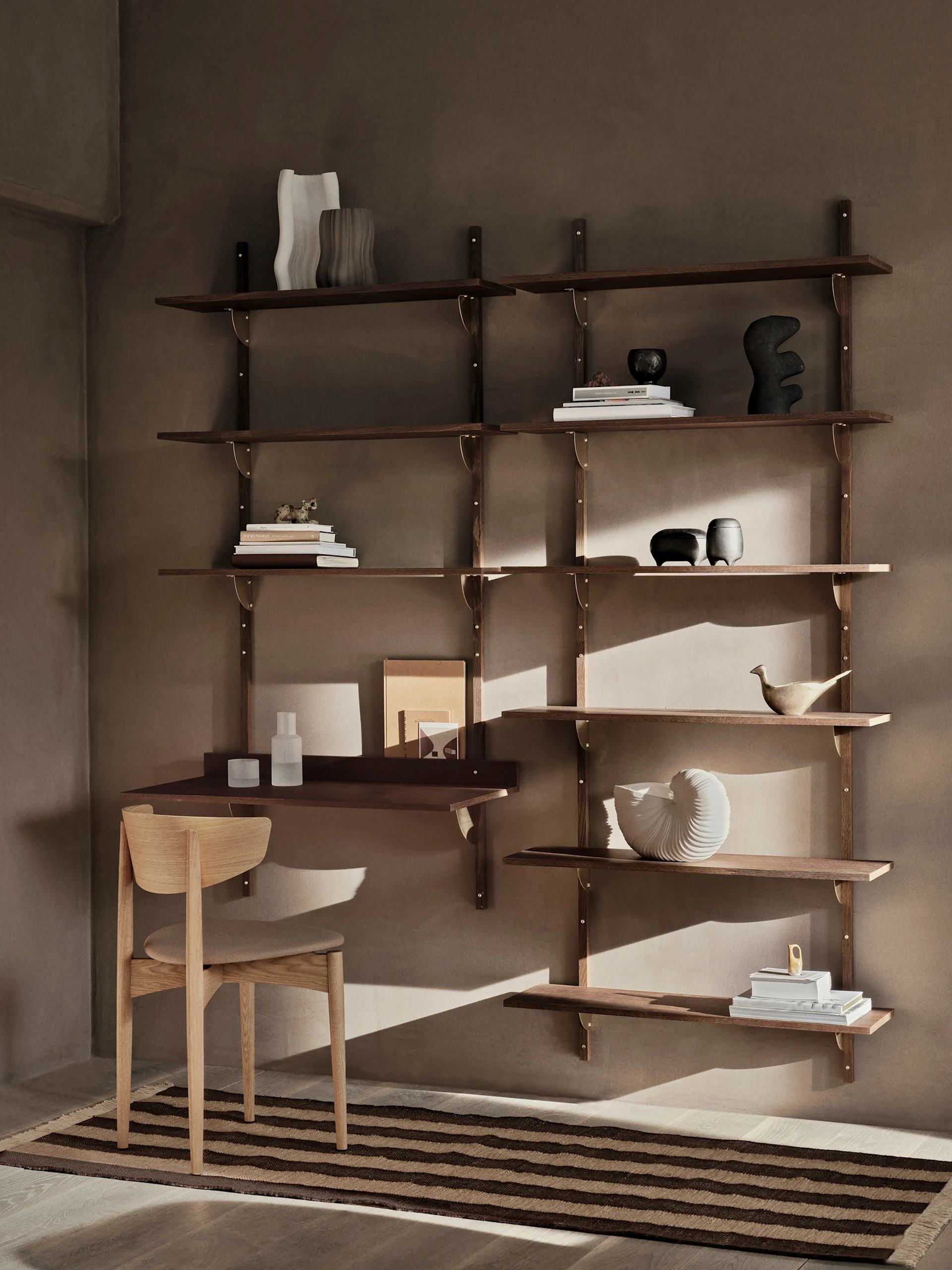 Une étagère murale moderne en bois foncé avec un bureau et une chaise, décorée de livres et d'objets, contre un mur marron chaud avec un tapis rayé.