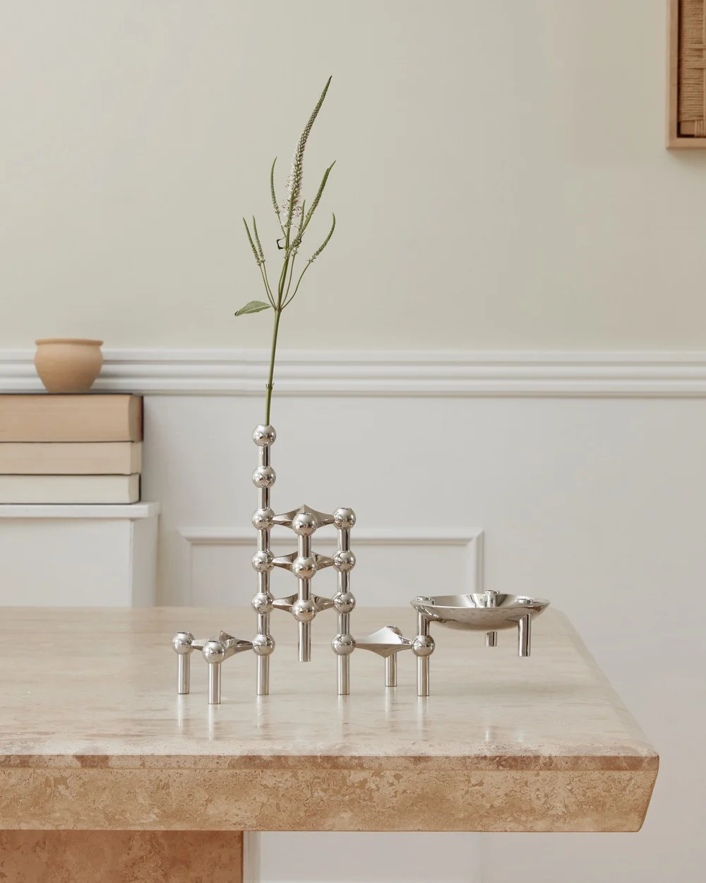 Un système de bougeoirs modulaires en argent poli avec une tige de plante et une petite coupelle sur une table en travertin.