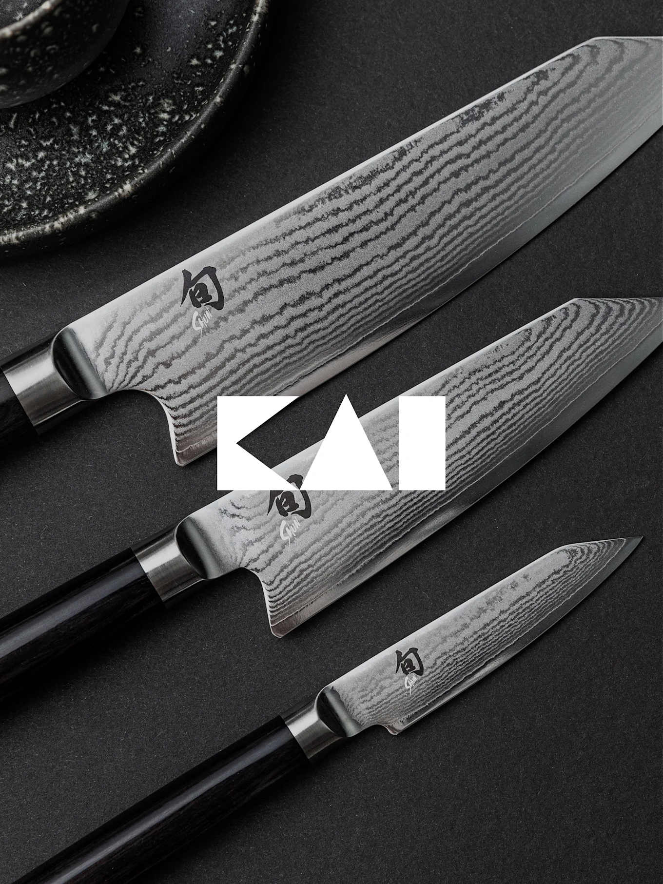 Trois couteaux de cuisine avec des lames en acier de Damas et des manches sombres, disposés en diagonale sur une surface sombre.