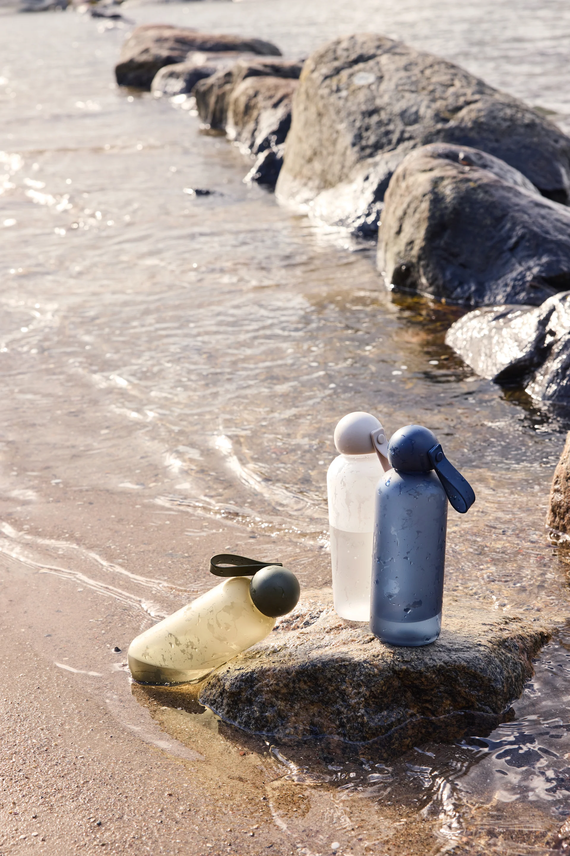 Trois gourdes réutilisables, de couleur jaune, blanc mat et bleu-gris, sont posées sur un rocher au bord de l'eau.