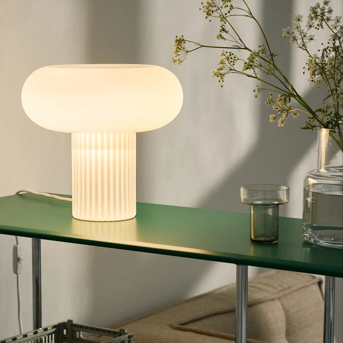 Lampe de table lumineuse en forme de champignon avec base nervurée sur une étagère vert foncé, avec des fleurs.
