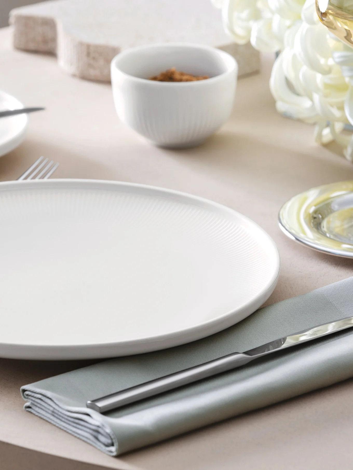 Une assiette creuse blanche côtelée, une serviette vert sauge et des couverts argentés sur une table avec un petit bol en arrière-plan.