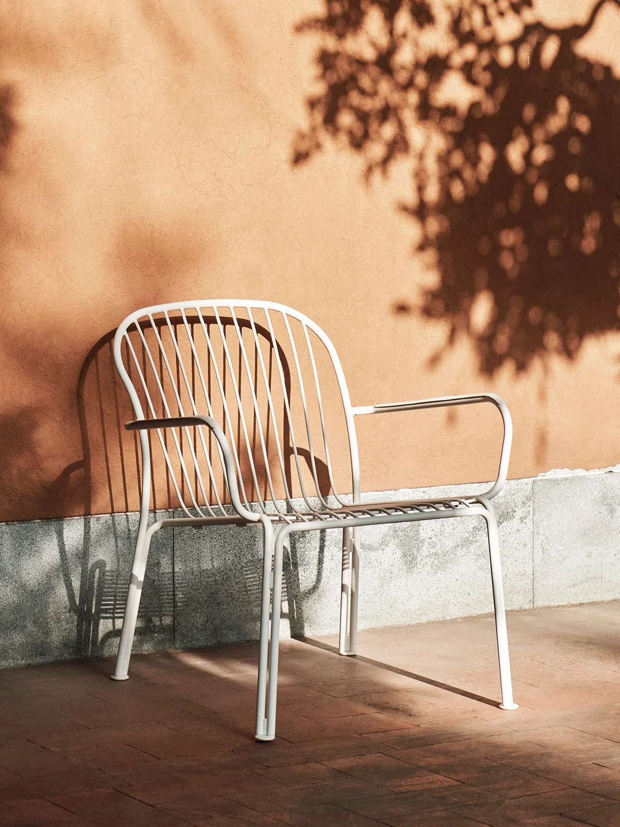 Chaise d'extérieur en métal blanc à lattes devant un mur orange avec des ombres d'arbres par une journée ensoleillée.