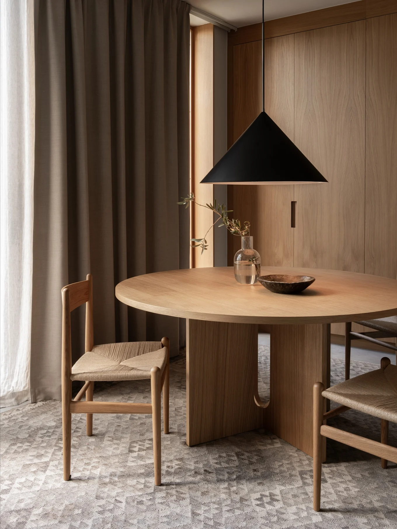 Un coin repas minimaliste avec une table ronde en bois, deux chaises aux assises tressées, une suspension noire et des murs en bois.