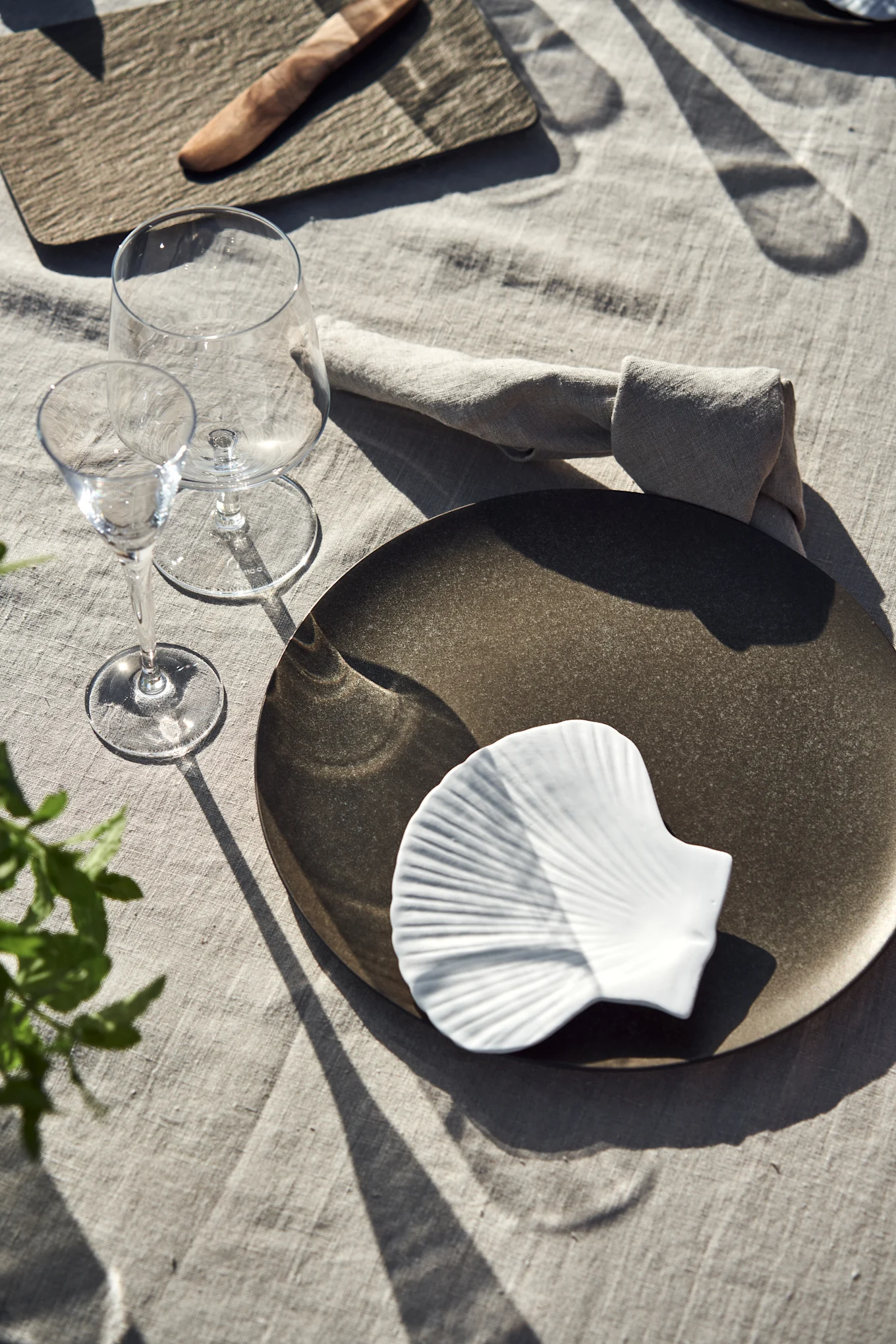 Un couvert individuel avec une assiette foncée, un plat blanc en forme de coquillage, deux verres à vin et une serviette en lin sur une nappe claire, avec des ombres marquées.