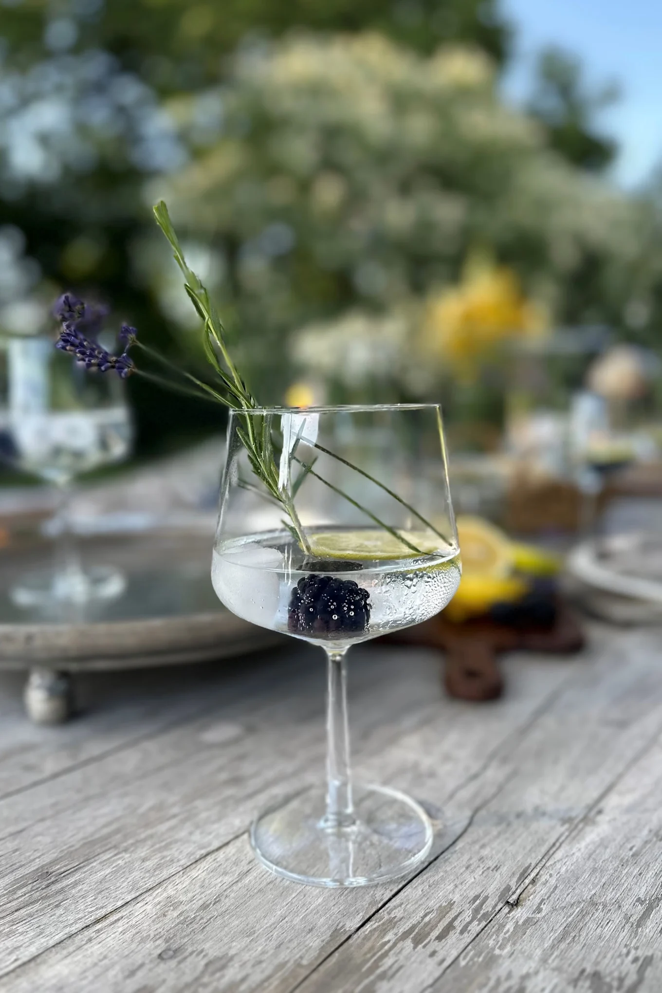 Un verre à pied transparent posé sur une table en bois, contenant une mûre, une rondelle de citron vert, une branche de lavande et un liquide transparent.
