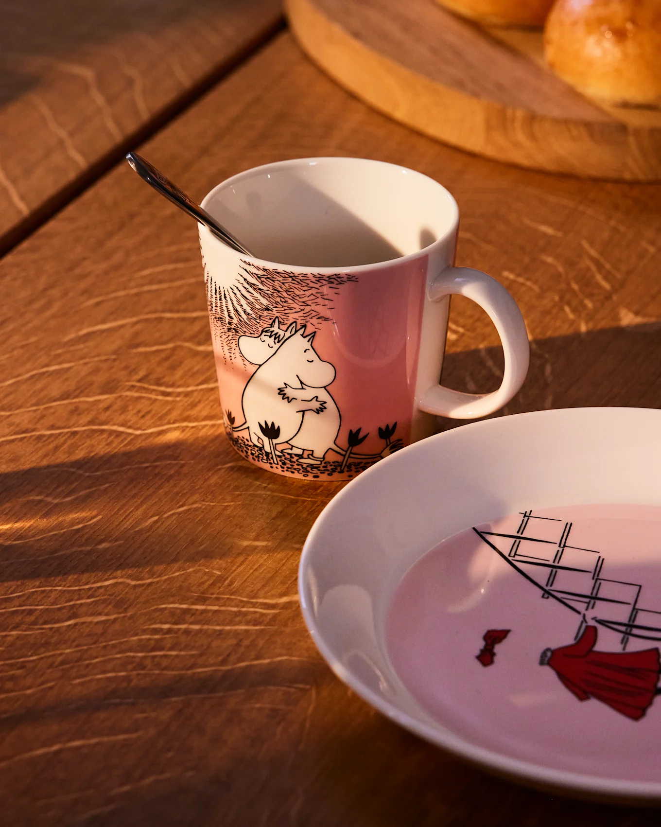 Tasse Moomin rose avec Moomintroll et Snorkmaiden, à côté d'une assiette assortie avec un voilier, sur une table en bois.