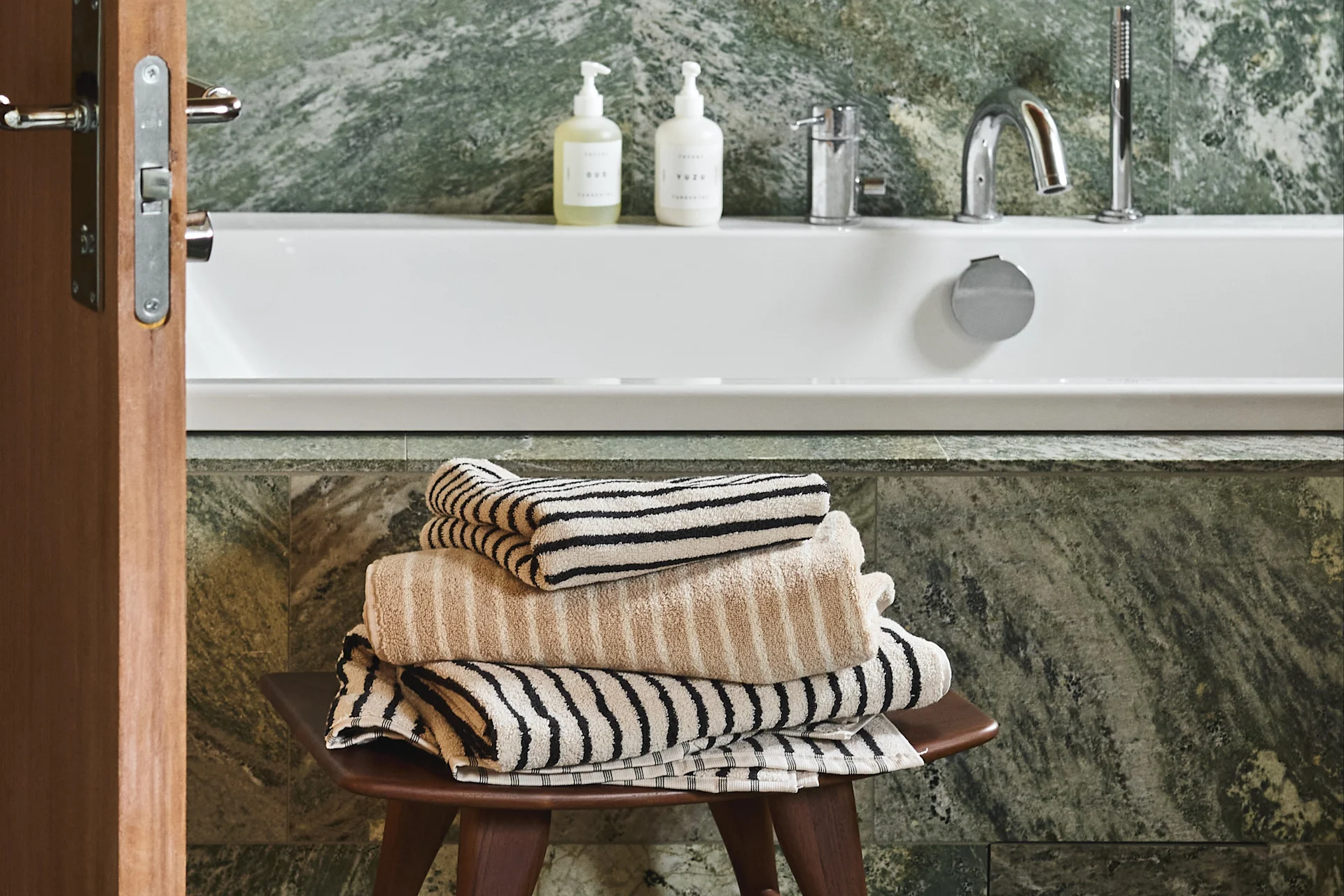 Une pile de serviettes beiges et rayées sur un tabouret en bois dans une salle de bain avec une baignoire blanche et des murs en marbre vert.