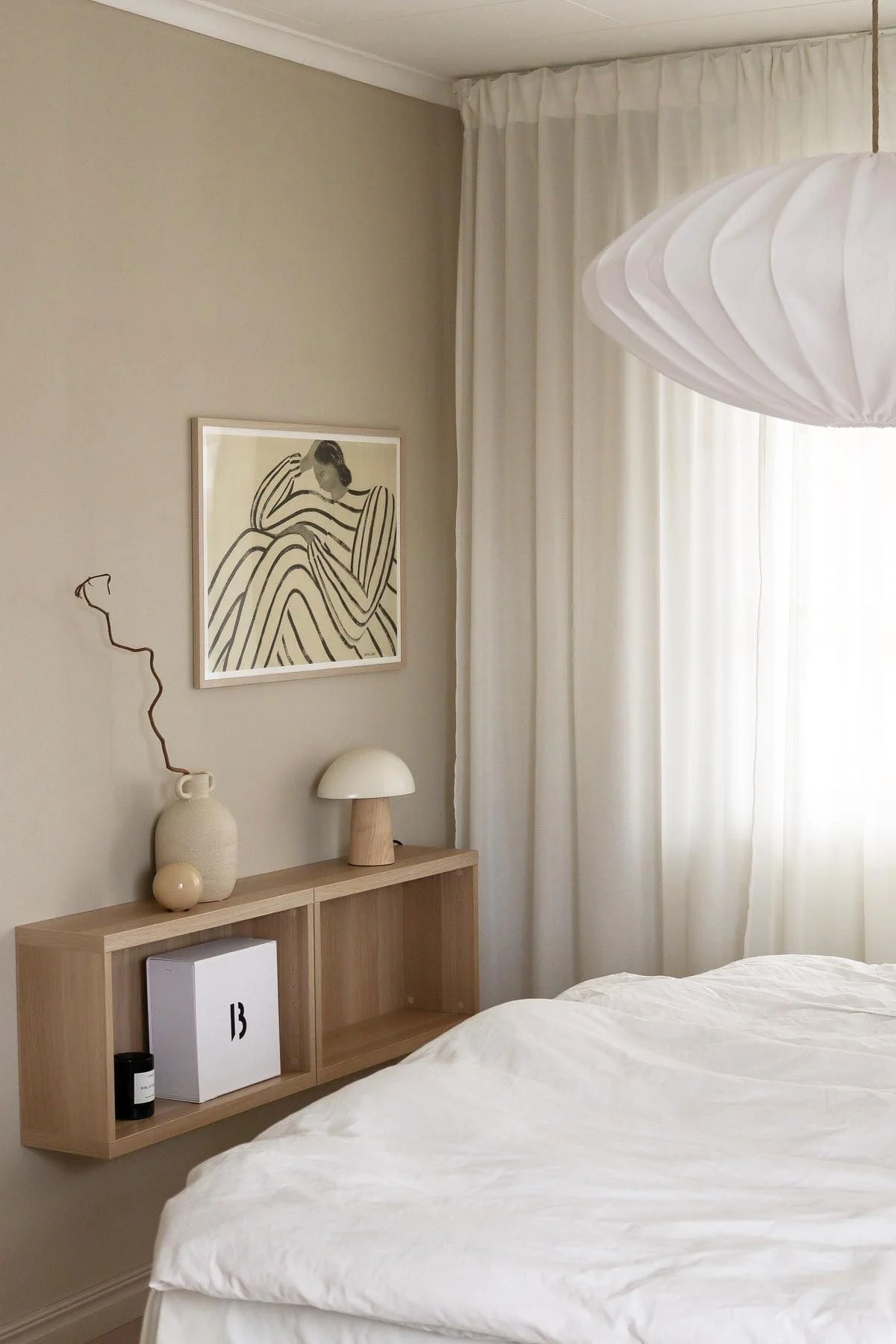 Chambre minimaliste avec literie blanche, étagère flottante en bois décorée, art abstrait, rideaux transparents et suspension plissée.
