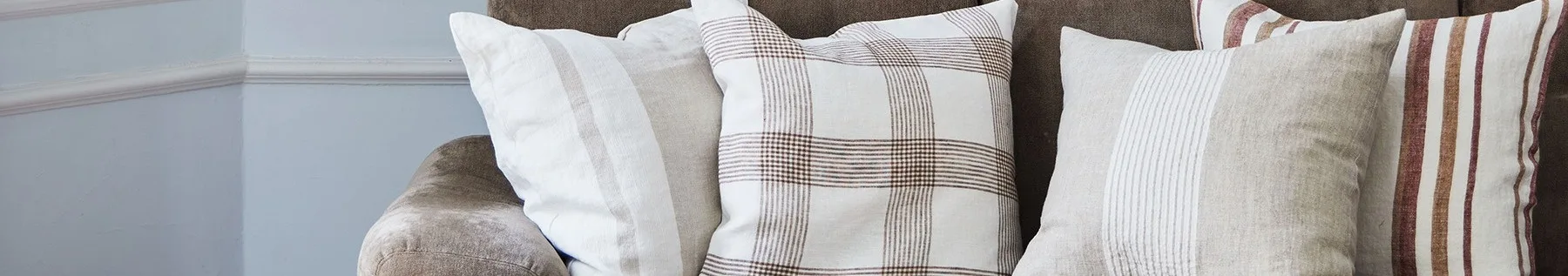 Gros plan de trois coussins sur un canapé marron, présentant des motifs blancs, beiges et à carreaux avec des rayures marron et rouges.