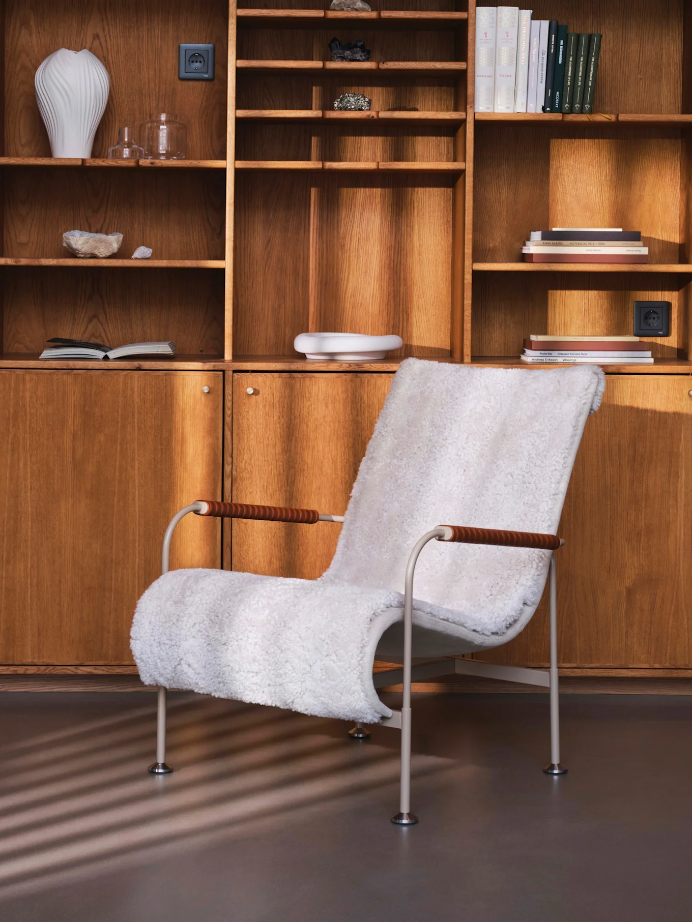 Fauteuil blanc en fausse fourrure devant une étagère murale en bois avec livres, vases et minéraux.