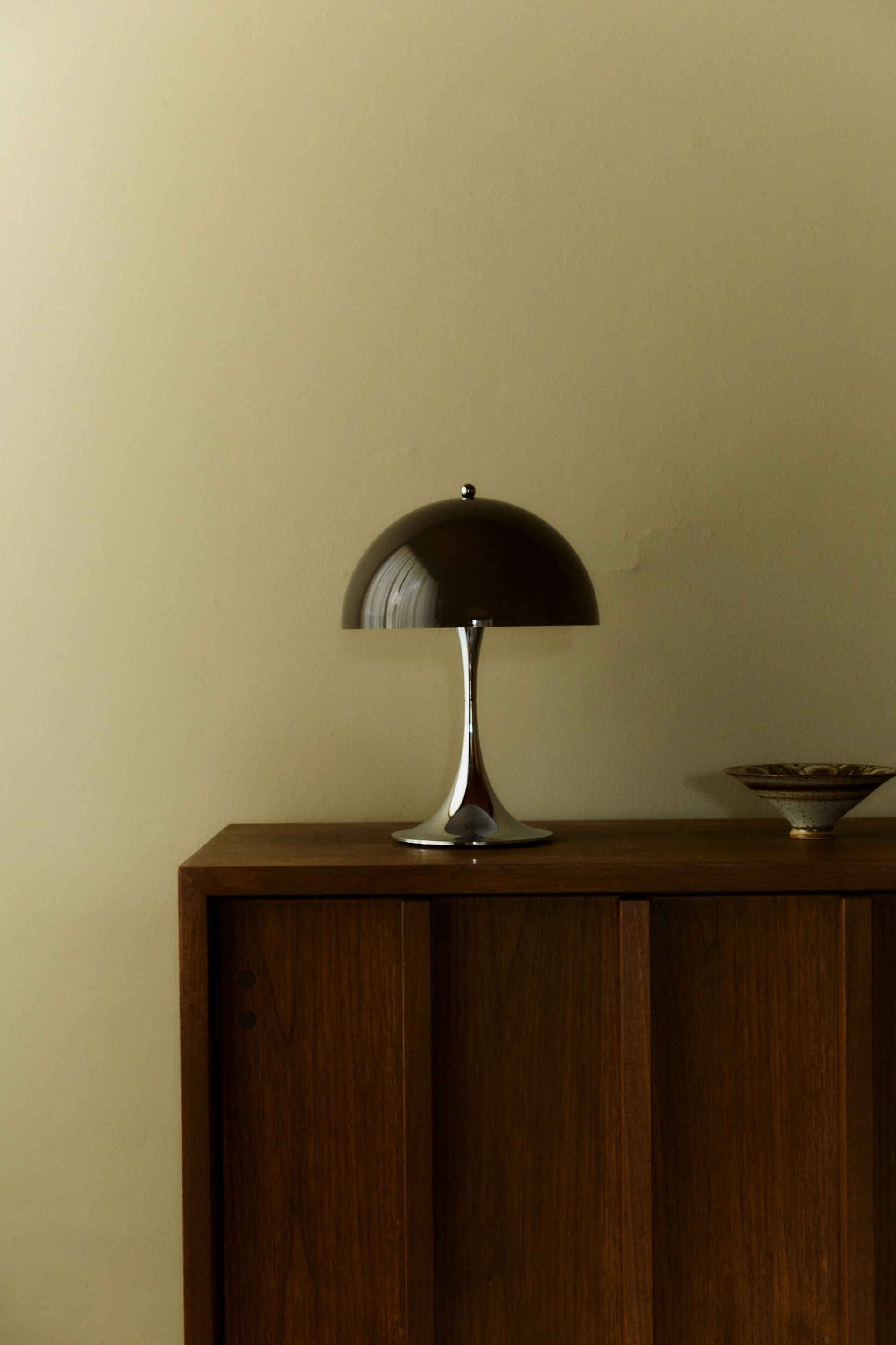 Une lampe de table champignon moderne et élégante avec un abat-jour en dôme foncé et une base chromée, posée sur une armoire en bois à côté d'un bol en céramique.