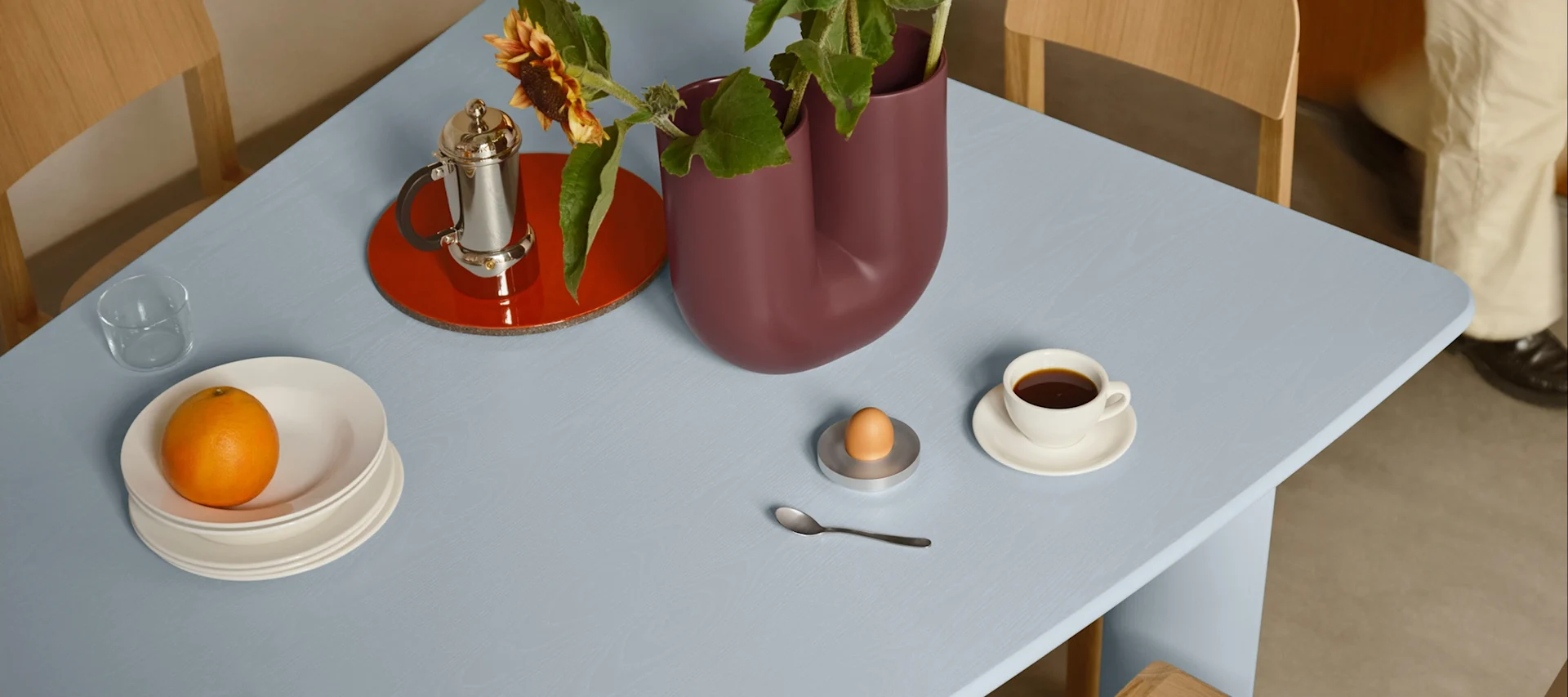 Table bleu clair dressée pour le petit-déjeuner: orange sur assiettes, café, œuf dans coquetier et vase avec tournesols.