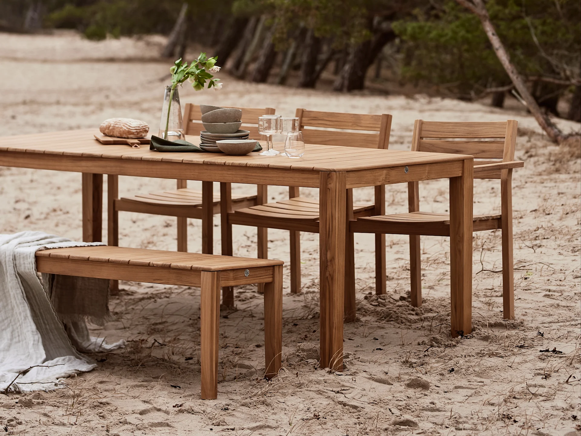 Ensemble repas extérieur en bois avec table, chaises et banc, décoré de vaisselle et d'une couverture sur une plage de sable.