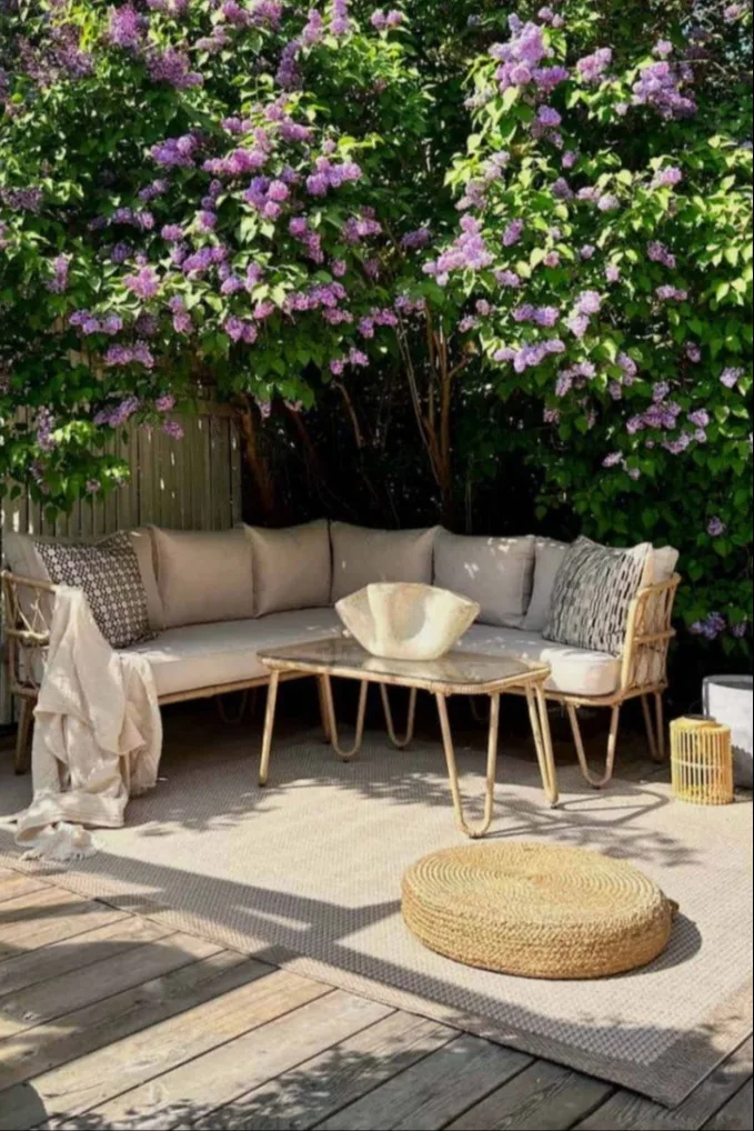 Patio extérieur avec canapé d'angle en osier, table basse et pouf sous un lilas en fleurs.