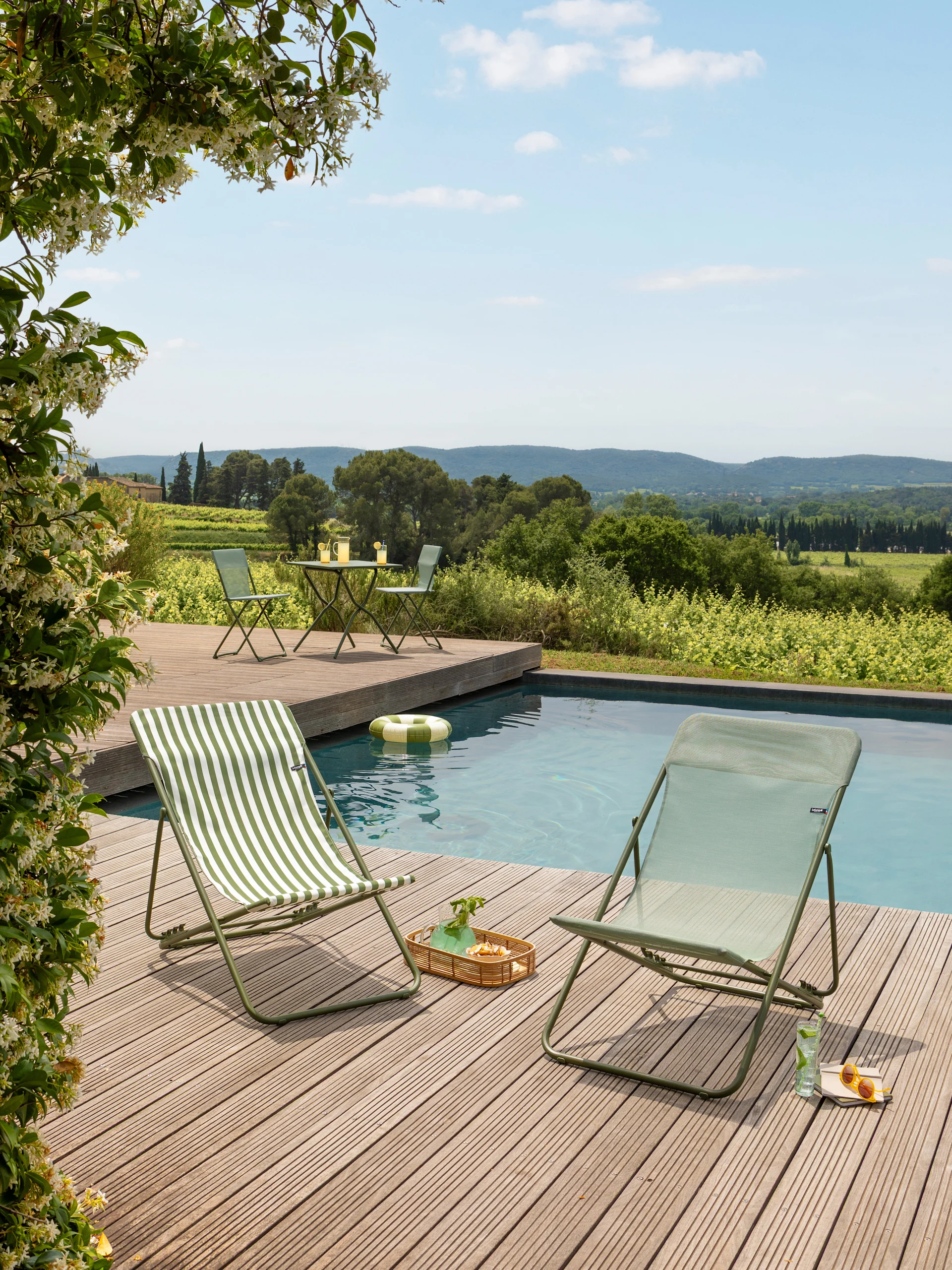 Deux chaises longues pliantes vertes, une rayée, sur une terrasse en bois près d'une piscine avec vue sur les vignobles.