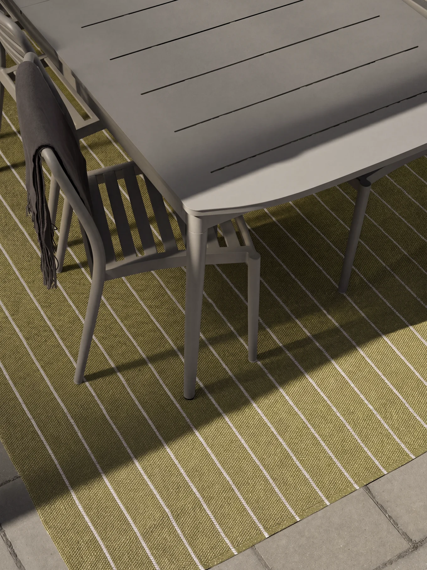 Table et chaises de salle à manger grises d'extérieur sur un tapis rayé vert et blanc. Une couverture gris foncé est drapée sur une chaise.