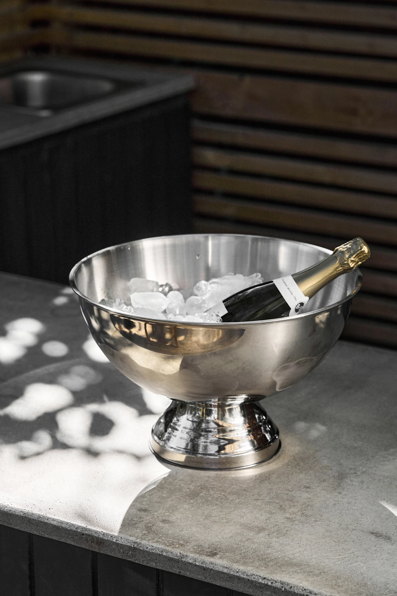 Une bouteille de champagne repose dans un seau à glace en acier inoxydable rempli de glaçons sur un comptoir en béton gris, avec des taches de soleil.