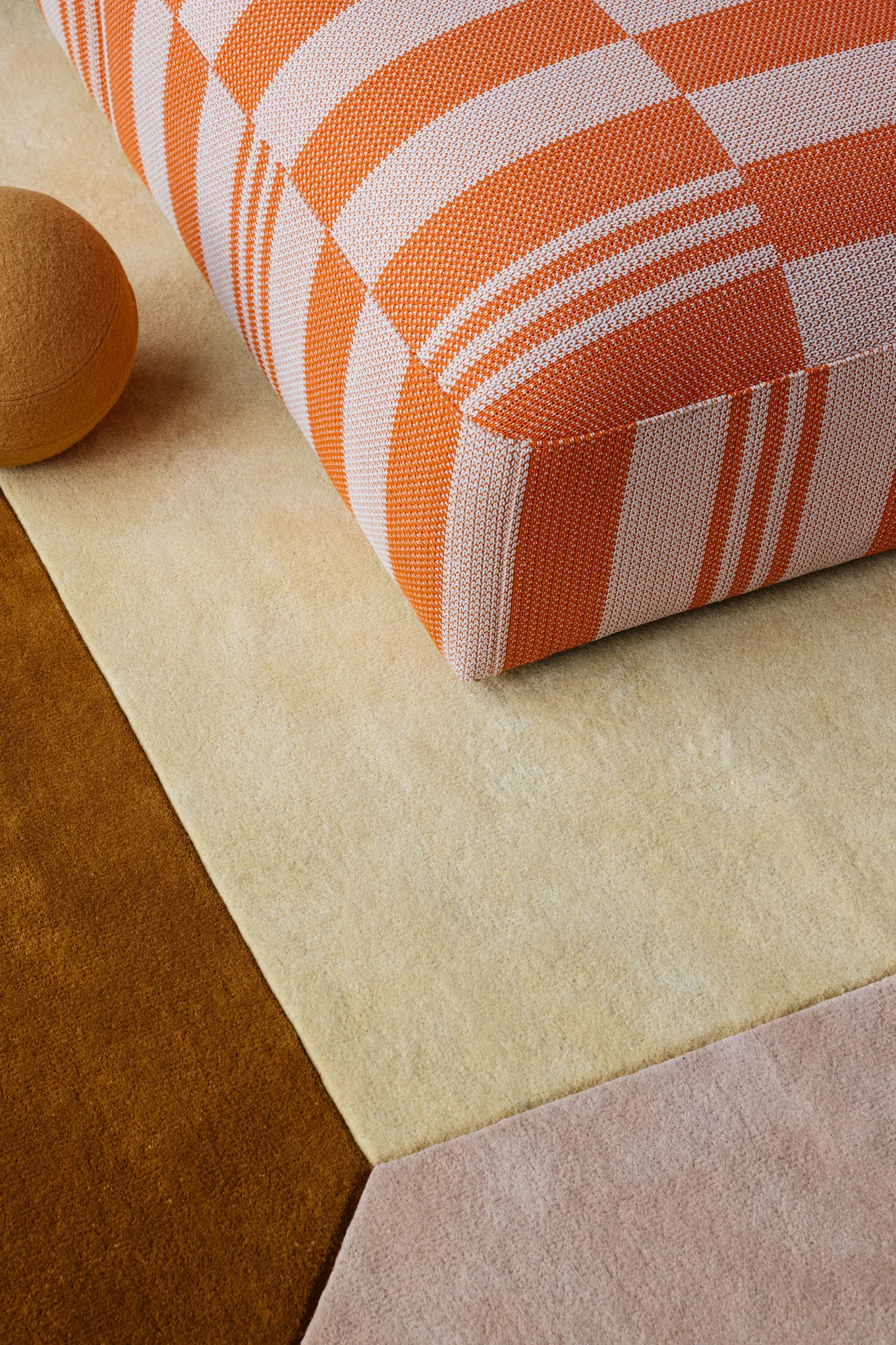 Un coussin à carreaux orange et blanc posé sur un tapis géométrique composé de sections beige clair, marron et rose. Une sphère marron se trouve à proximité.