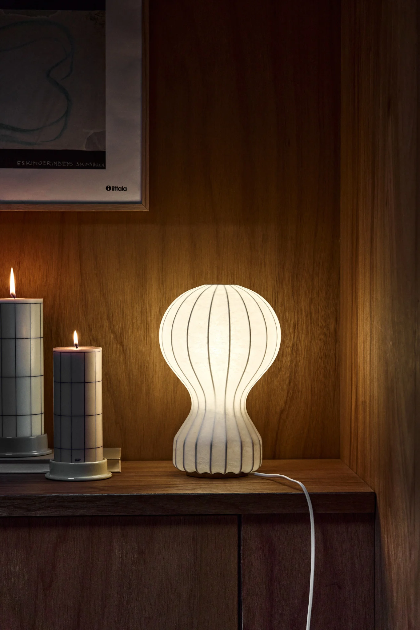 Lampe de table blanche côtelée éclairée, deux bougies à motif quadrillé et art encadré sur étagère en bois.