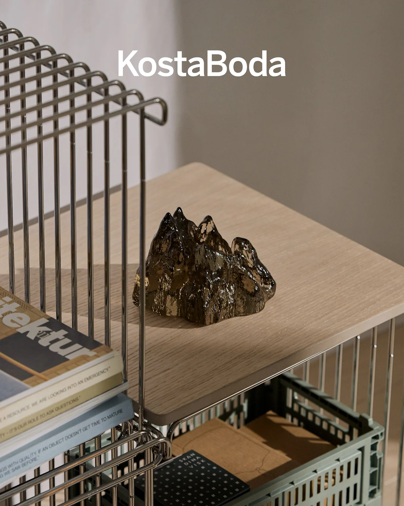 Nom de la marque KostaBoda au-dessus d'une sculpture en verre sombre, transparente, en forme de montagne sur une étagère en bois clair.