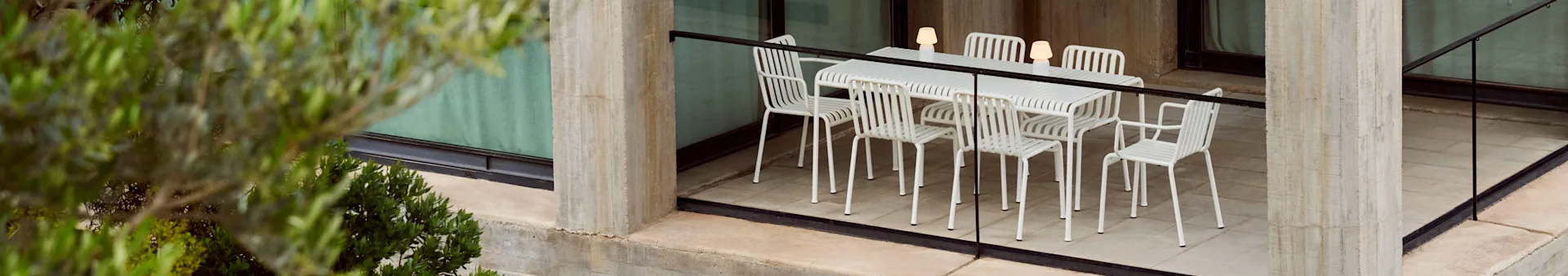 Un ensemble de salle à manger d'extérieur avec une table blanche à lattes et six chaises assorties, éclairé par trois lampes portables, sur une terrasse en béton avec des plantes.