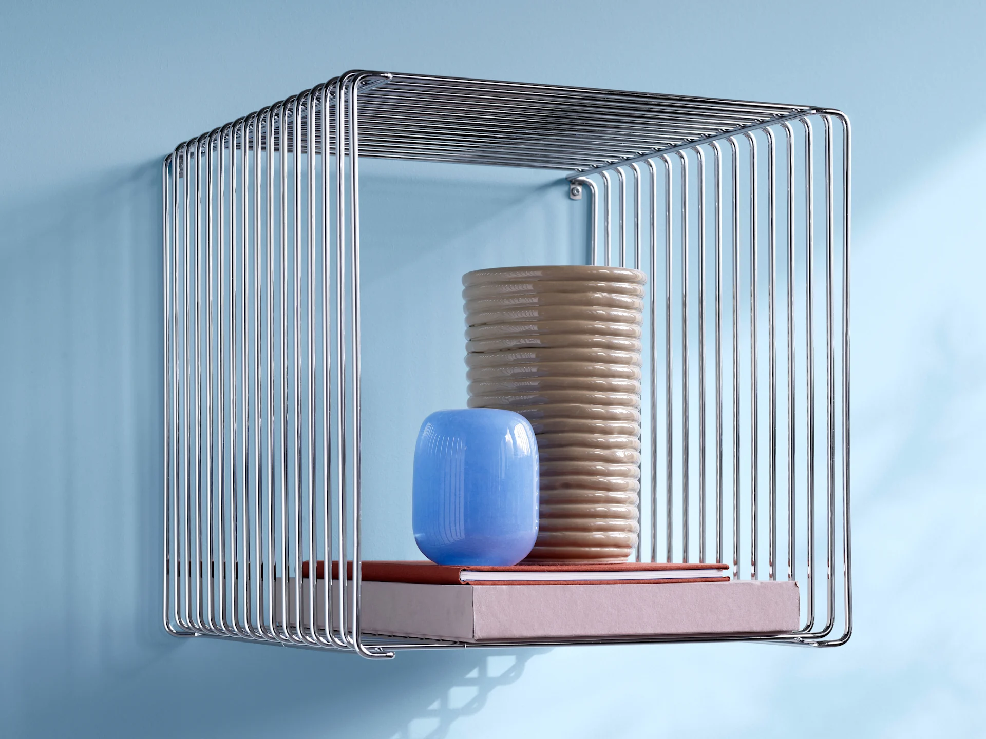 Une étagère en fil de chrome avec un grand vase beige nervuré, un petit vase bleu et deux livres sur un mur bleu clair.