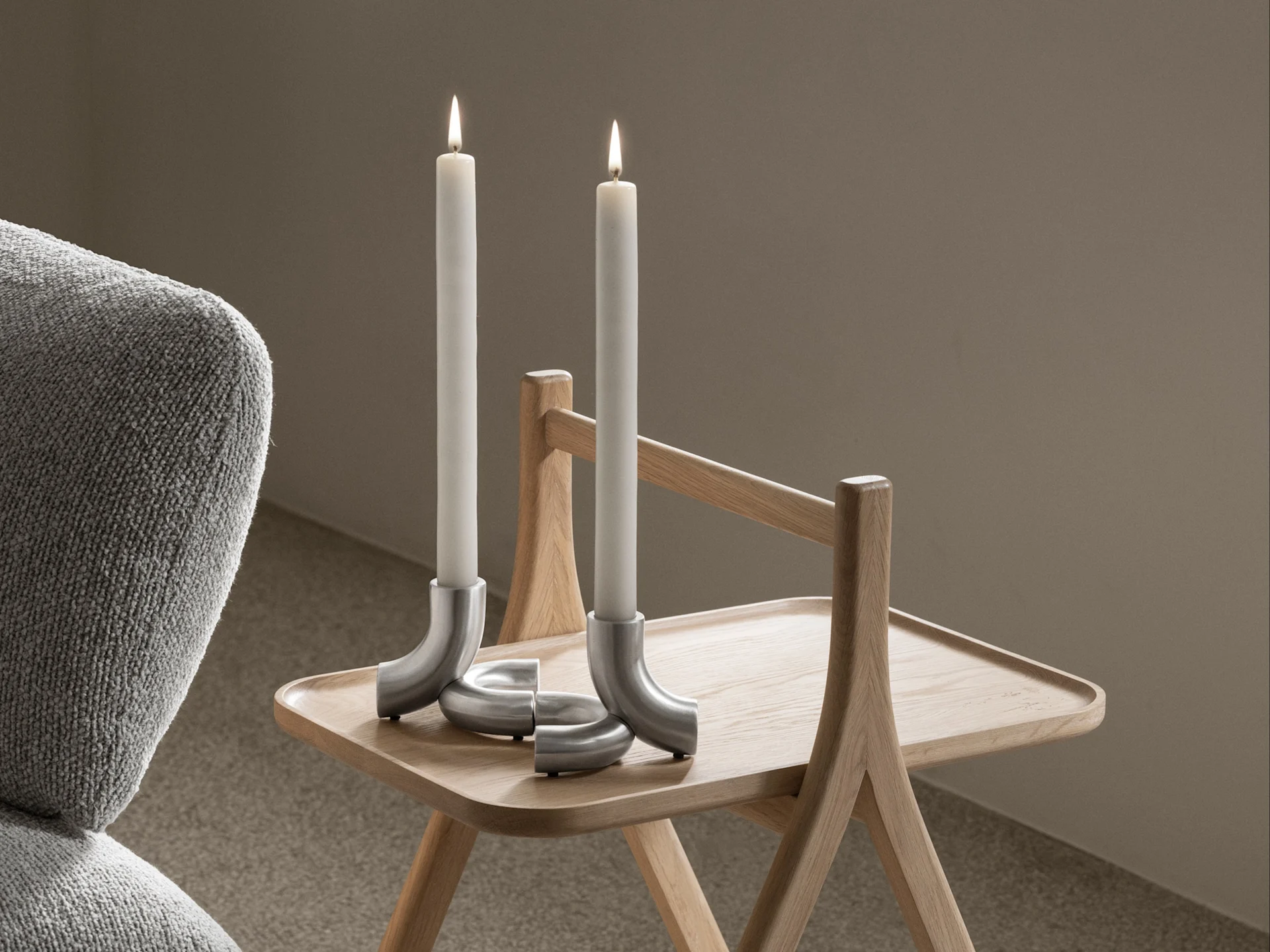 Deux bougies blanches allumées dans des bougeoirs argentés courbés sur une table d'appoint en bois clair, avec un fauteuil gris à gauche.