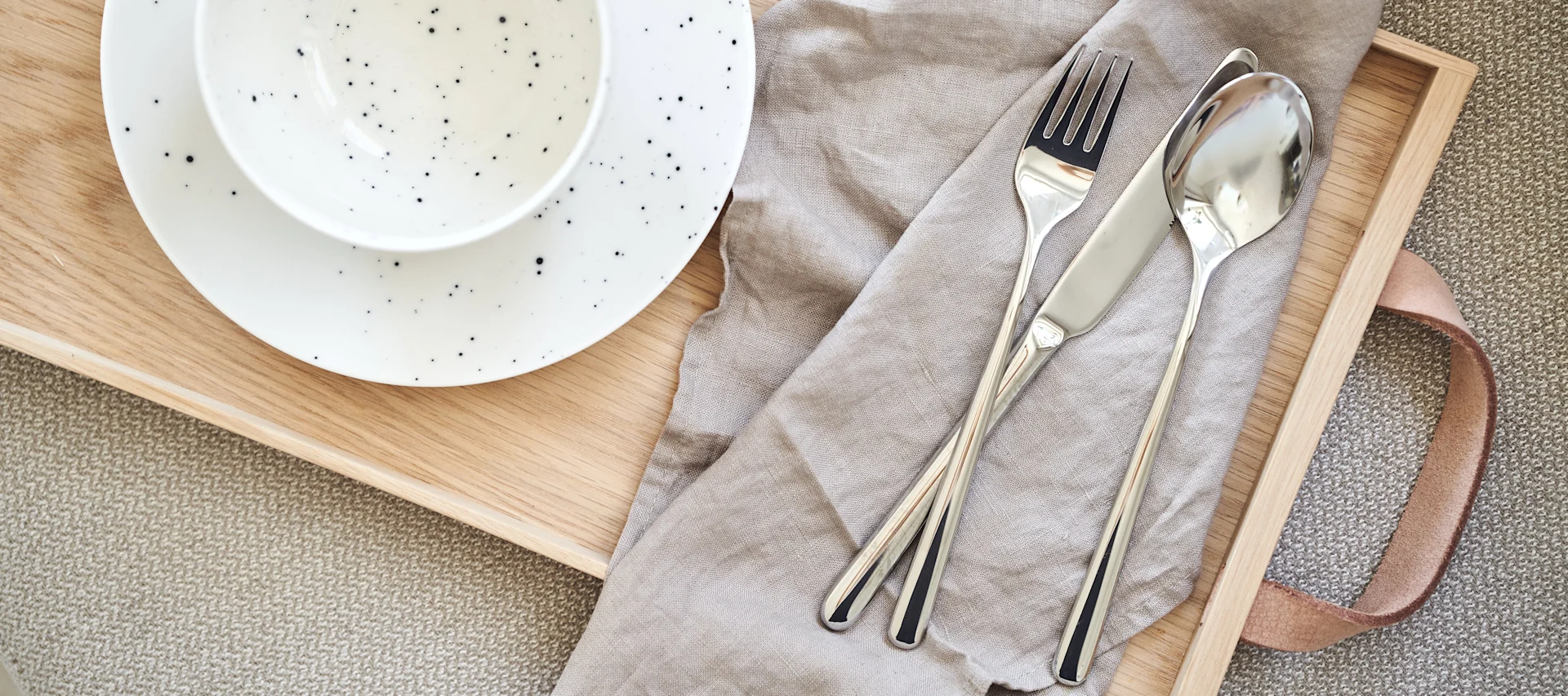 Service de table avec assiette et bol blancs mouchetés sur un plateau en bois, à côté d'une fourchette, d'un couteau et d'une cuillère sur une serviette en lin beige.