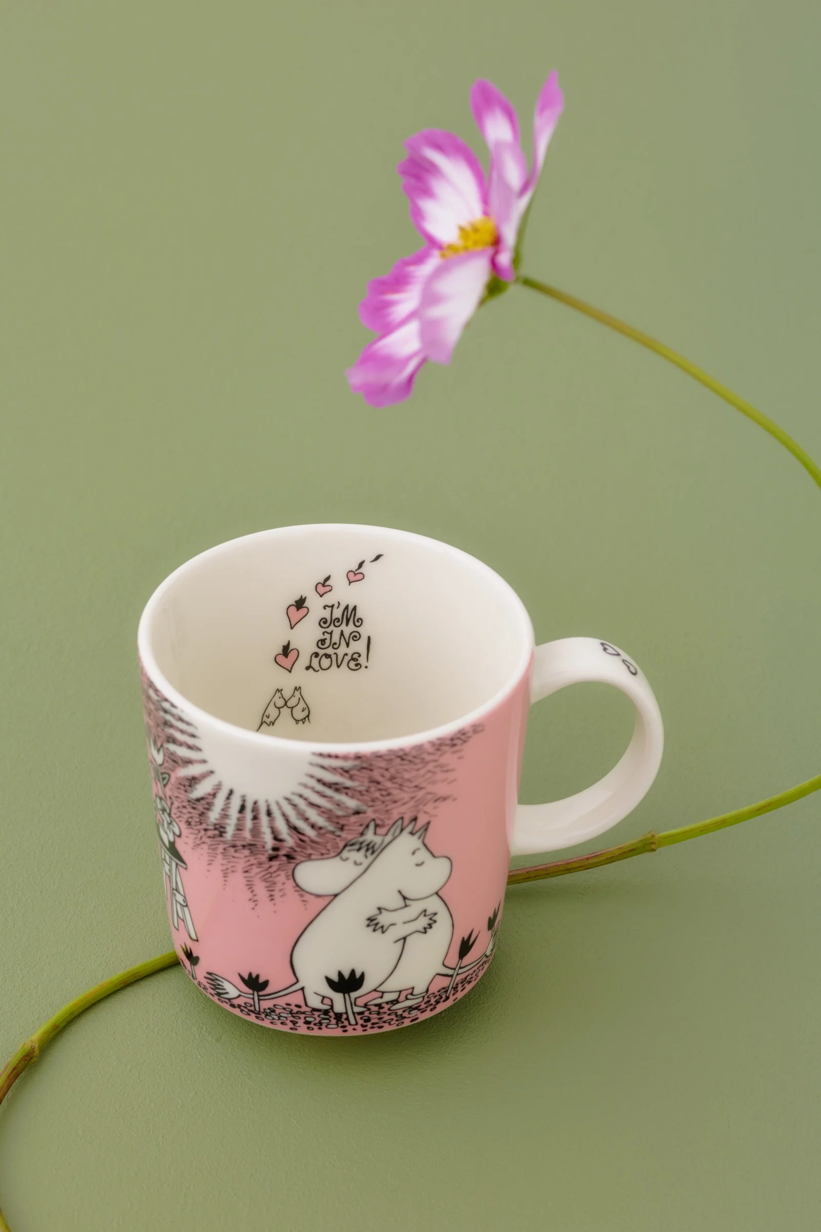 Tasse Moomin rose et blanche représentant Moomintroll et Snorkmaiden, avec l'inscription « I'M IN LOVE ! » à l'intérieur, sur fond vert orné d'une fleur de cosmos.