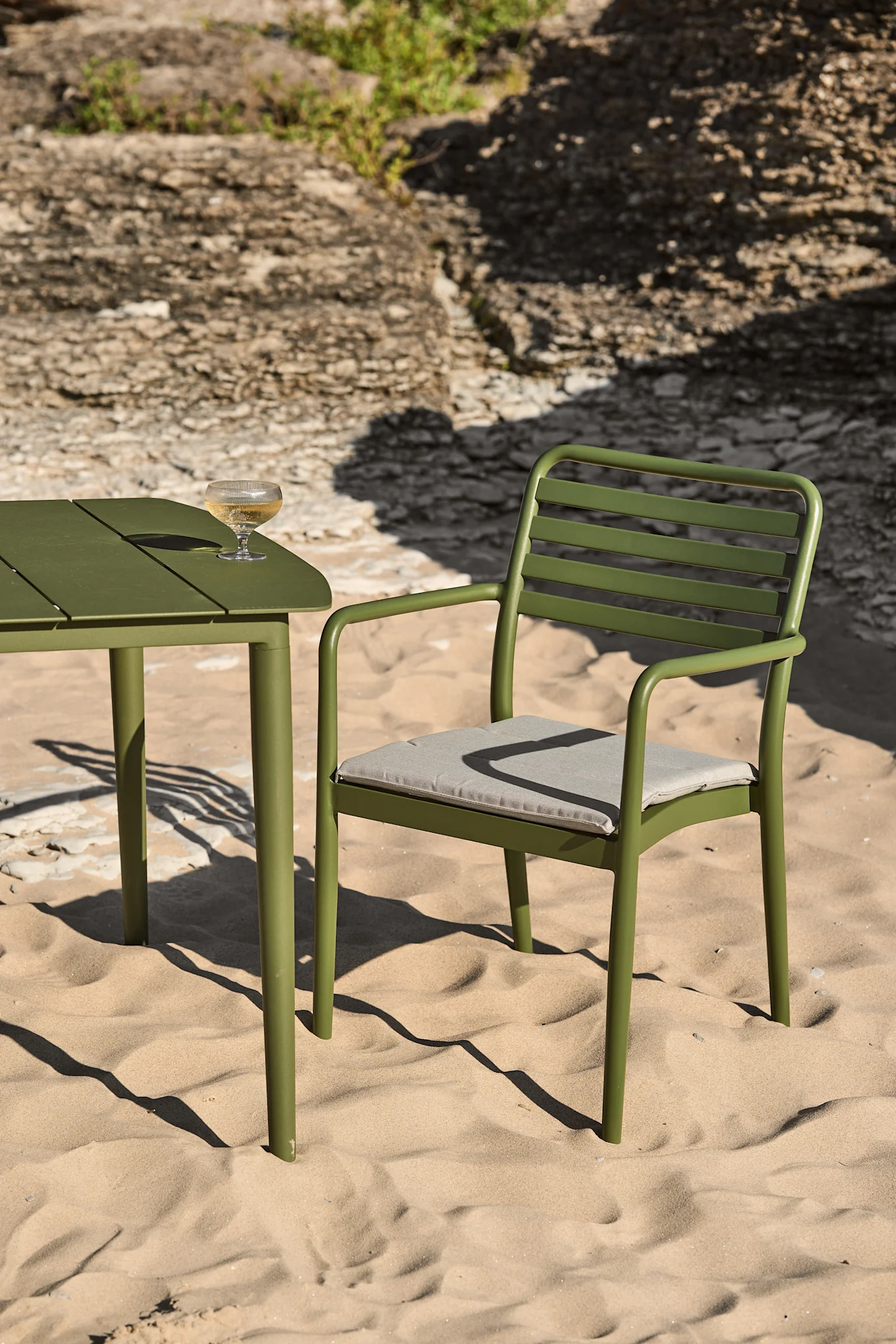 Une table et des chaises d'extérieur en métal vert sur une plage de sable, avec un verre de vin blanc posé sur la table.