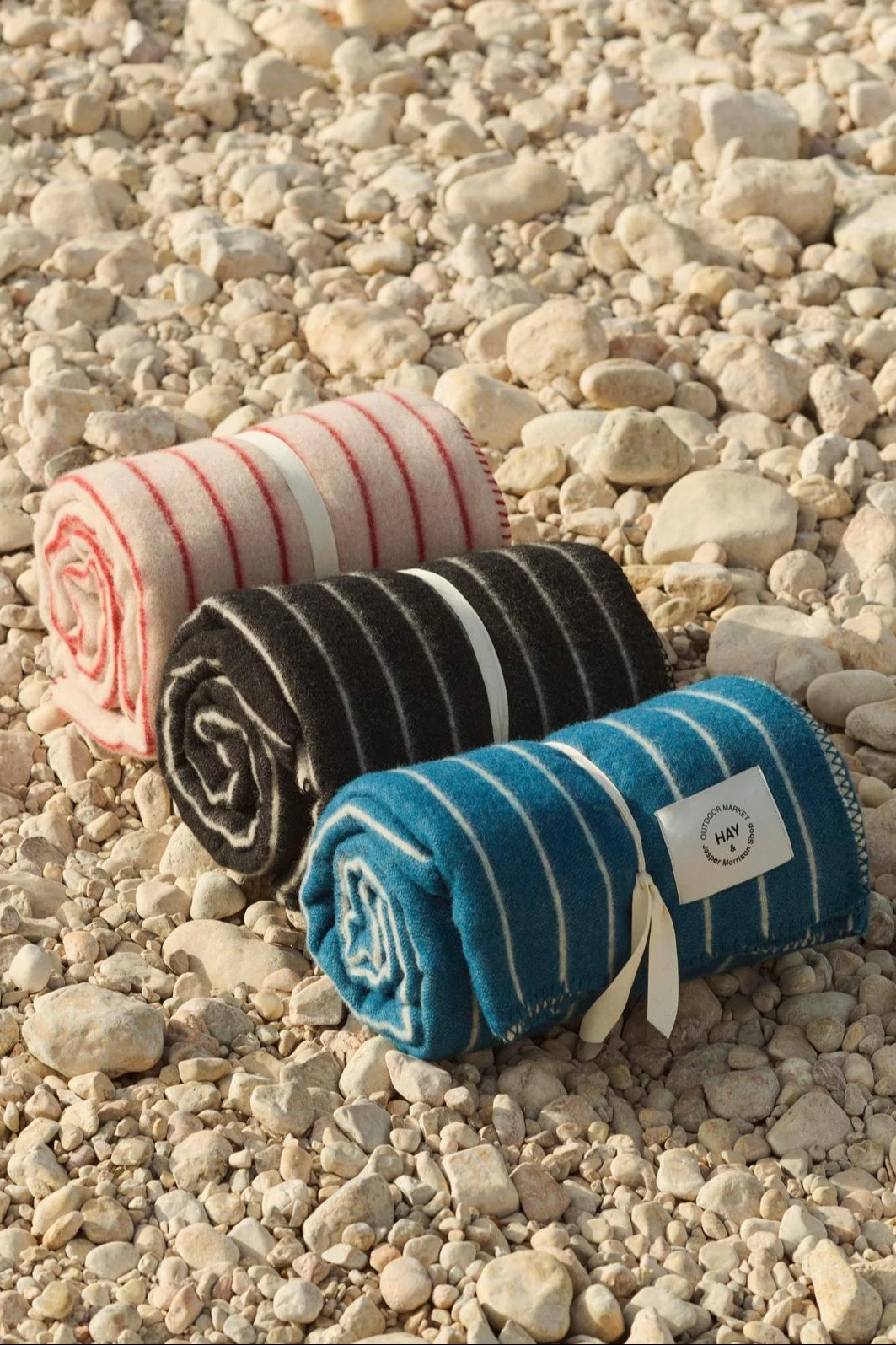 Trois couvertures rayées roulées en rose, noir et bleu, nouées avec des rubans sur une plage de galets.