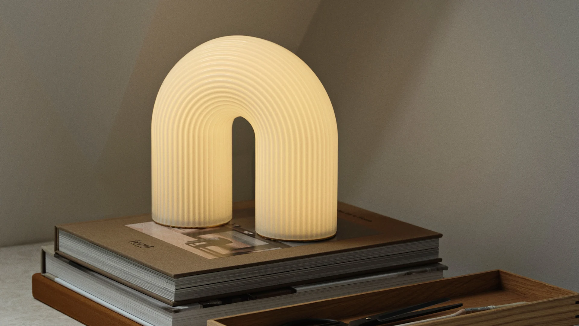 Lampe de table en forme d'arche, lumineuse et chaleureuse, avec une texture nervurée, posée sur une pile de livres marron, avec un plateau en bois à proximité.