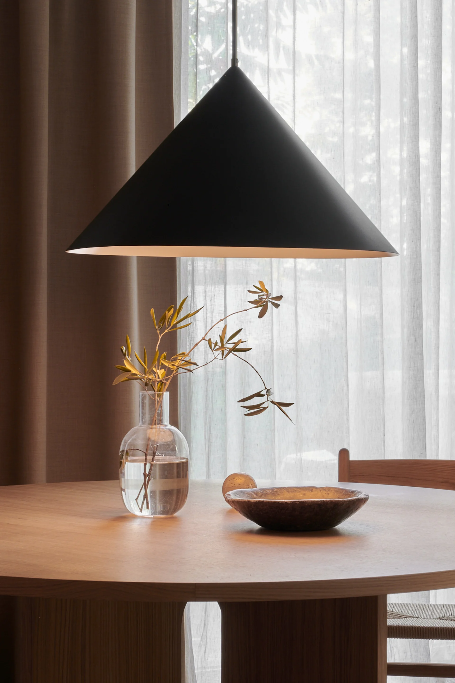 Table à manger ronde en bois avec une suspension conique noire au-dessus, présentant un vase transparent avec des branches et un bol en bois foncé.