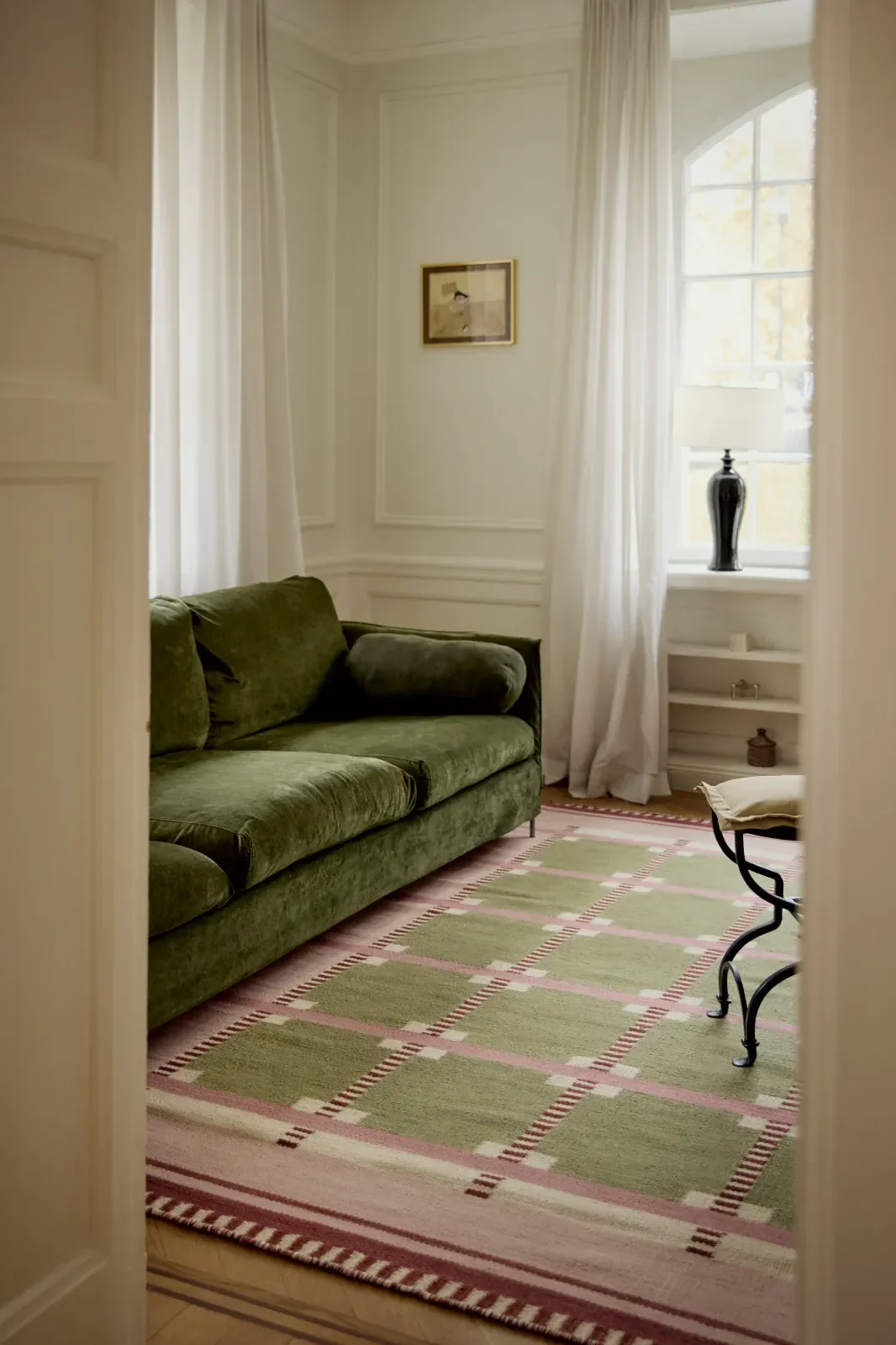 Un salon avec un canapé en velours vert, un tapis à motifs verts et roses et une fenêtre ornée de rideaux blancs.