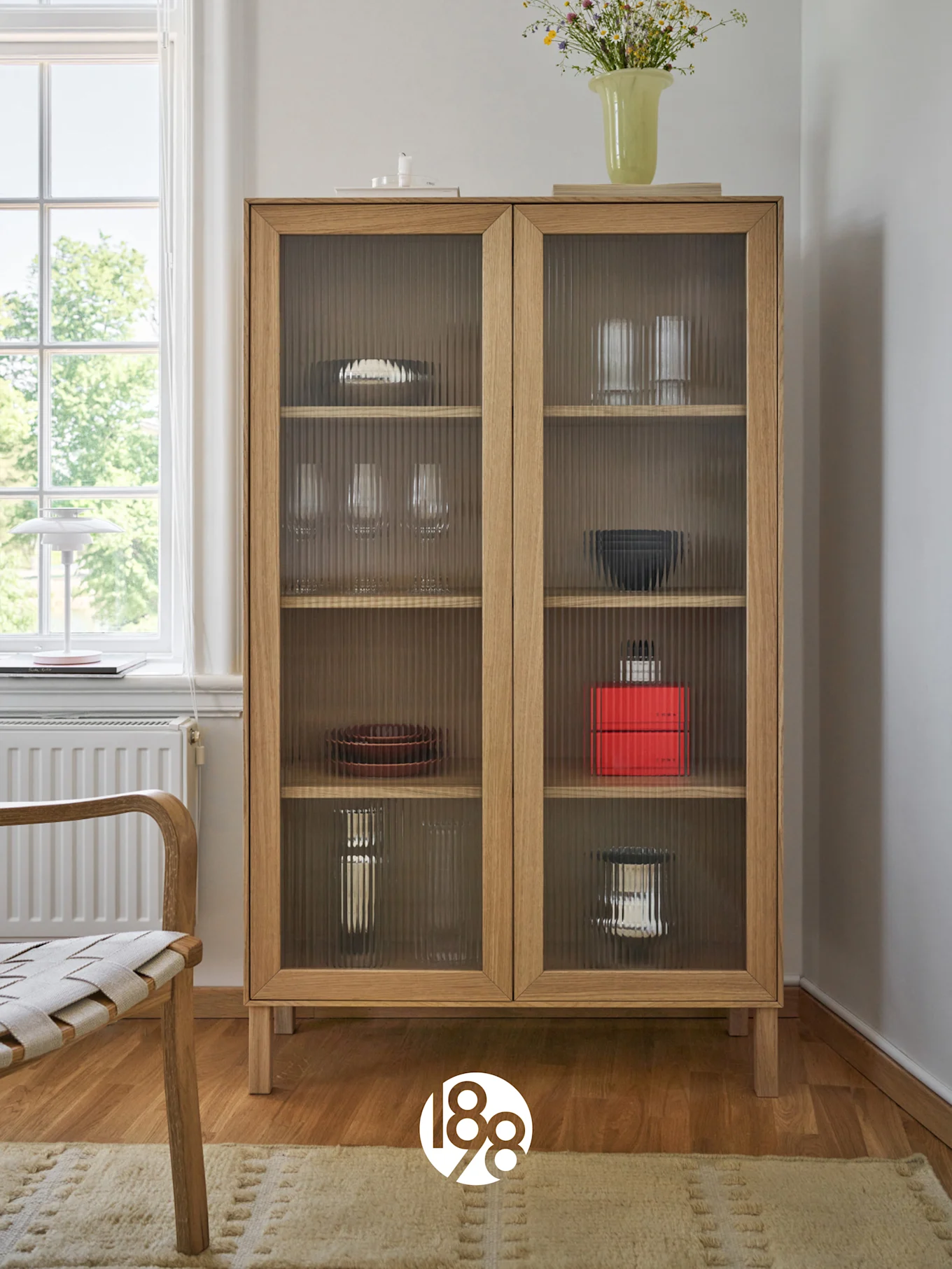 Une armoire en bois clair avec des portes en verre cannelé présentant de la vaisselle et des verres, surmontée d'un vase de fleurs sauvages. Une chaise et une fenêtre sont à gauche.