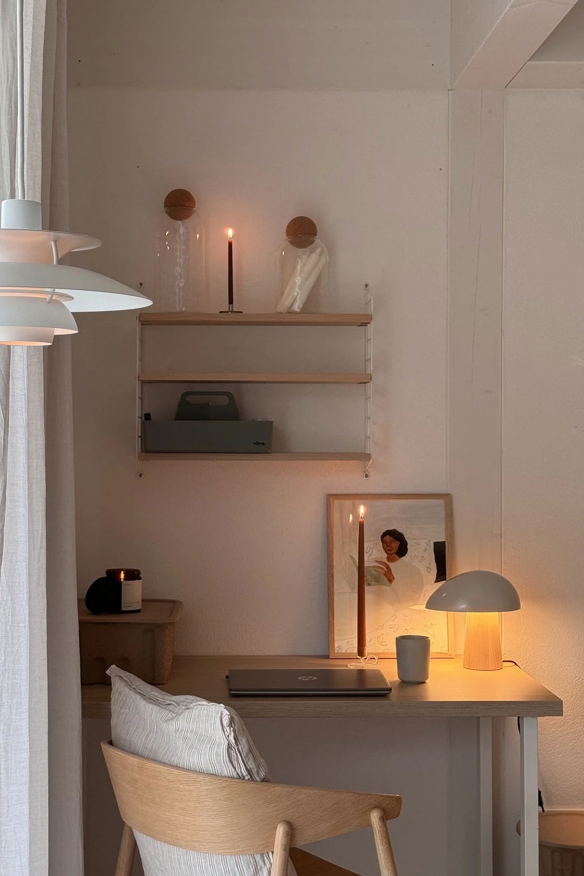Un bureau confortable avec un éclairage chaleureux, un ordinateur portable fermé, deux bougies allumées, une lampe moderne et une œuvre d'art encadrée.