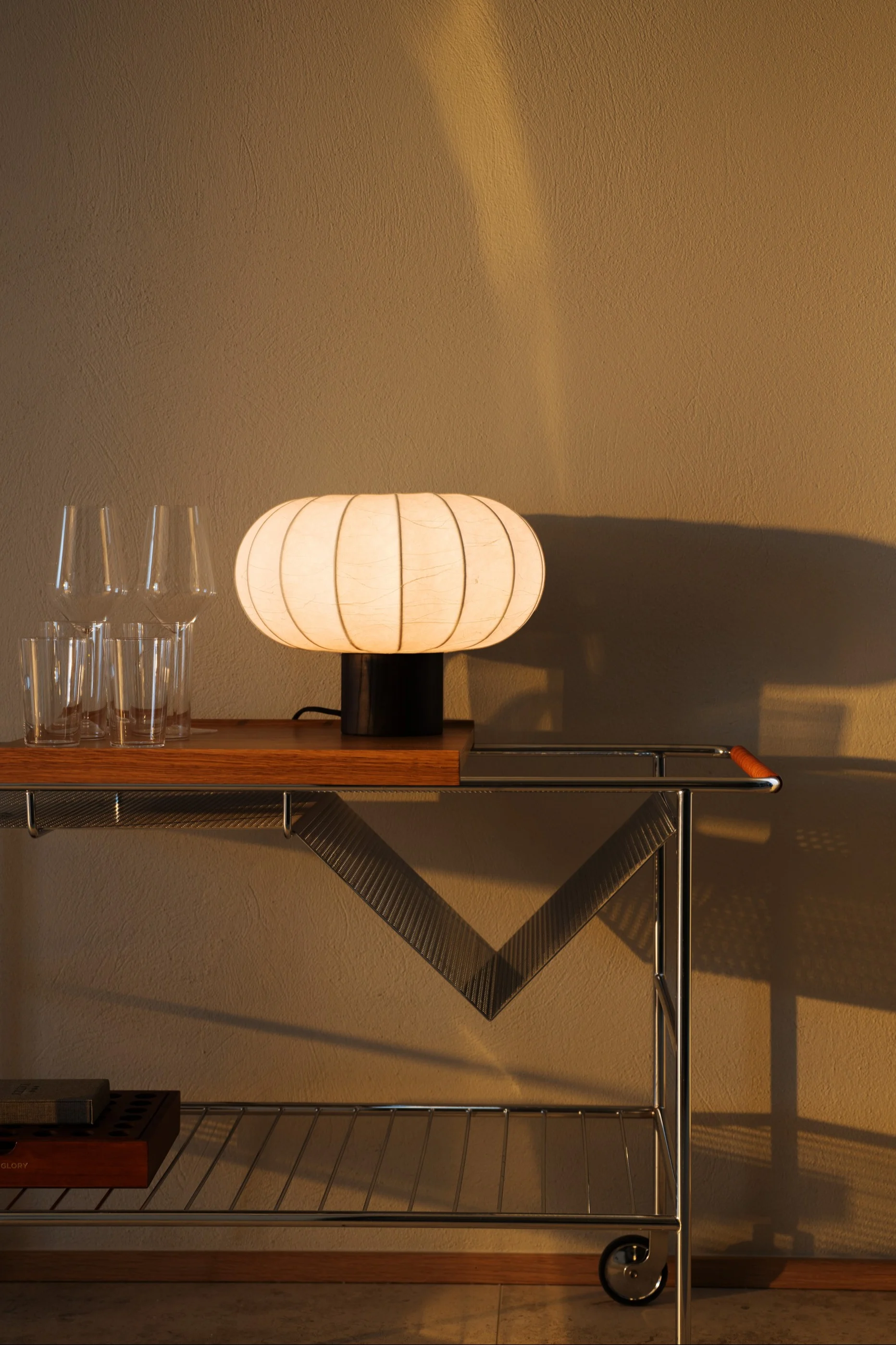 Une lampe ovale lumineuse et côtelée repose sur un chariot de bar en bois et chrome à côté de verres clairs, sur un mur texturé en arrière-plan.
