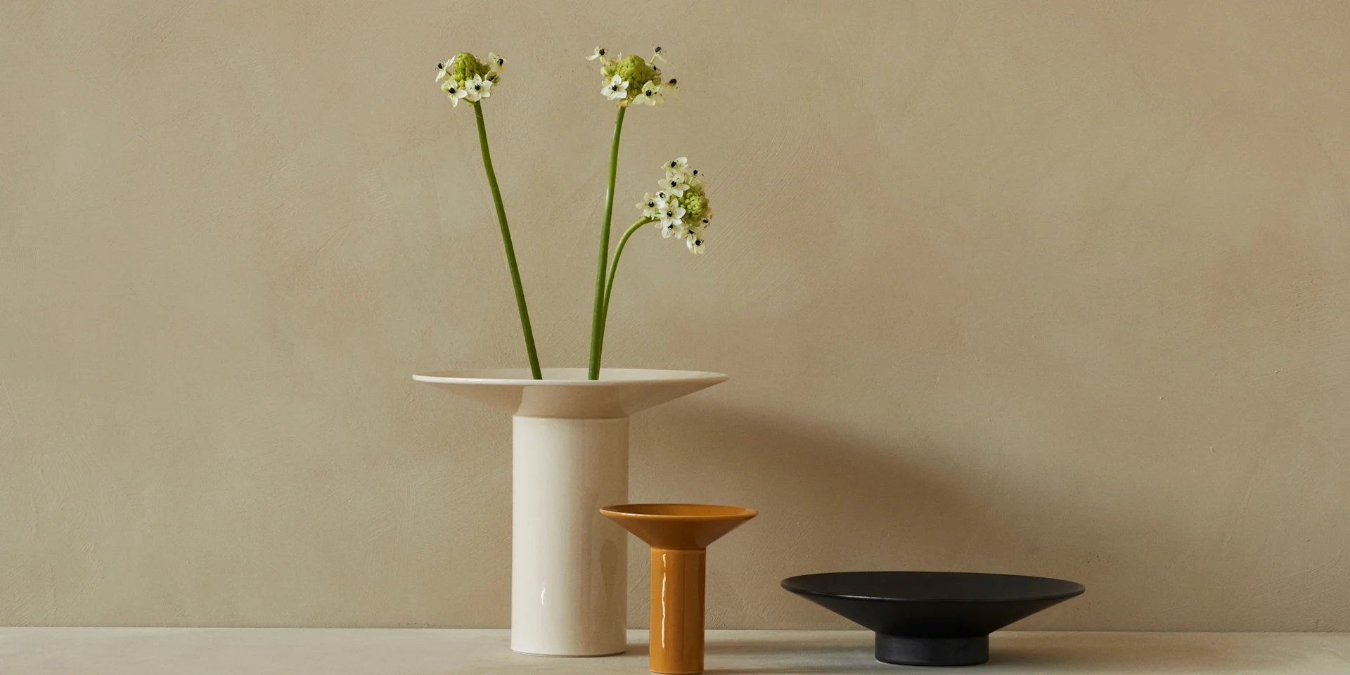 Trois vases en céramique blancs, ambrés et noirs disposés contre un mur beige texturé, avec des fleurs d'Ornithogalum blanches dans le vase blanc.