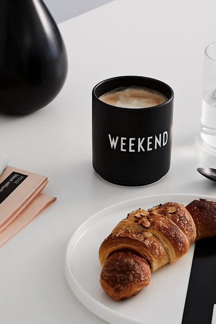 Mug à café : Le mug noir avec l’inscription « Weekend » de Design Letters est posé sur une table, devant lui une assiette avec un croissant.