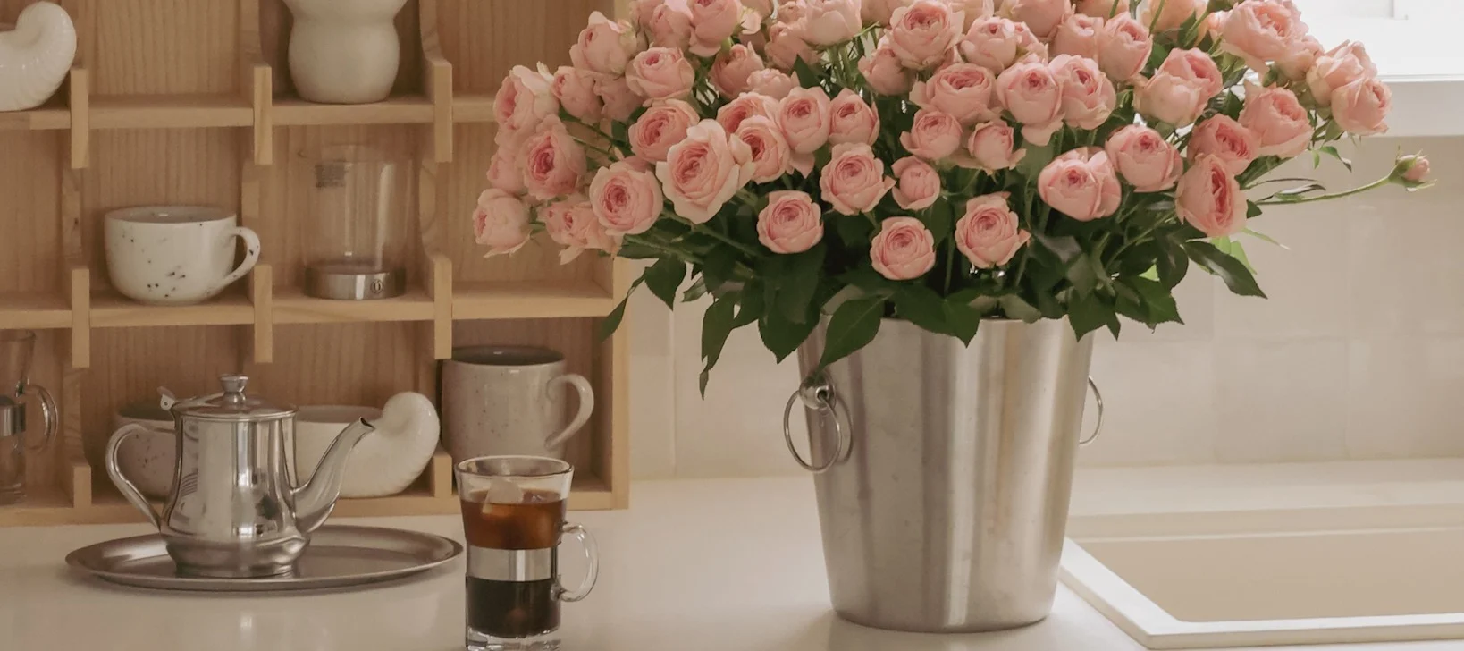 Sur un buffet de cuisine se trouvent une cafetière, une tasse de café et un grand bouquet de roses roses dans un seau à champagne Nordwik - un cadeau attentionné pour la Saint-Valentin.