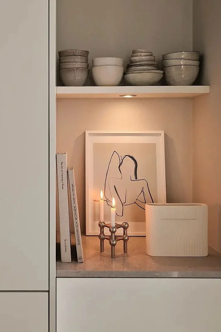Une étagère chaleureuse où se trouvent des bols en céramique, des livres, des tableaux encadrés, des bougies allumées dans un bougeoir en laiton et un vase strié.