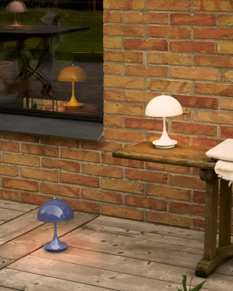 Trois lampes en forme de champignon en blanc, bleu et jaune sur une terrasse en bois près d'un mur de briques et d'une fenêtre.